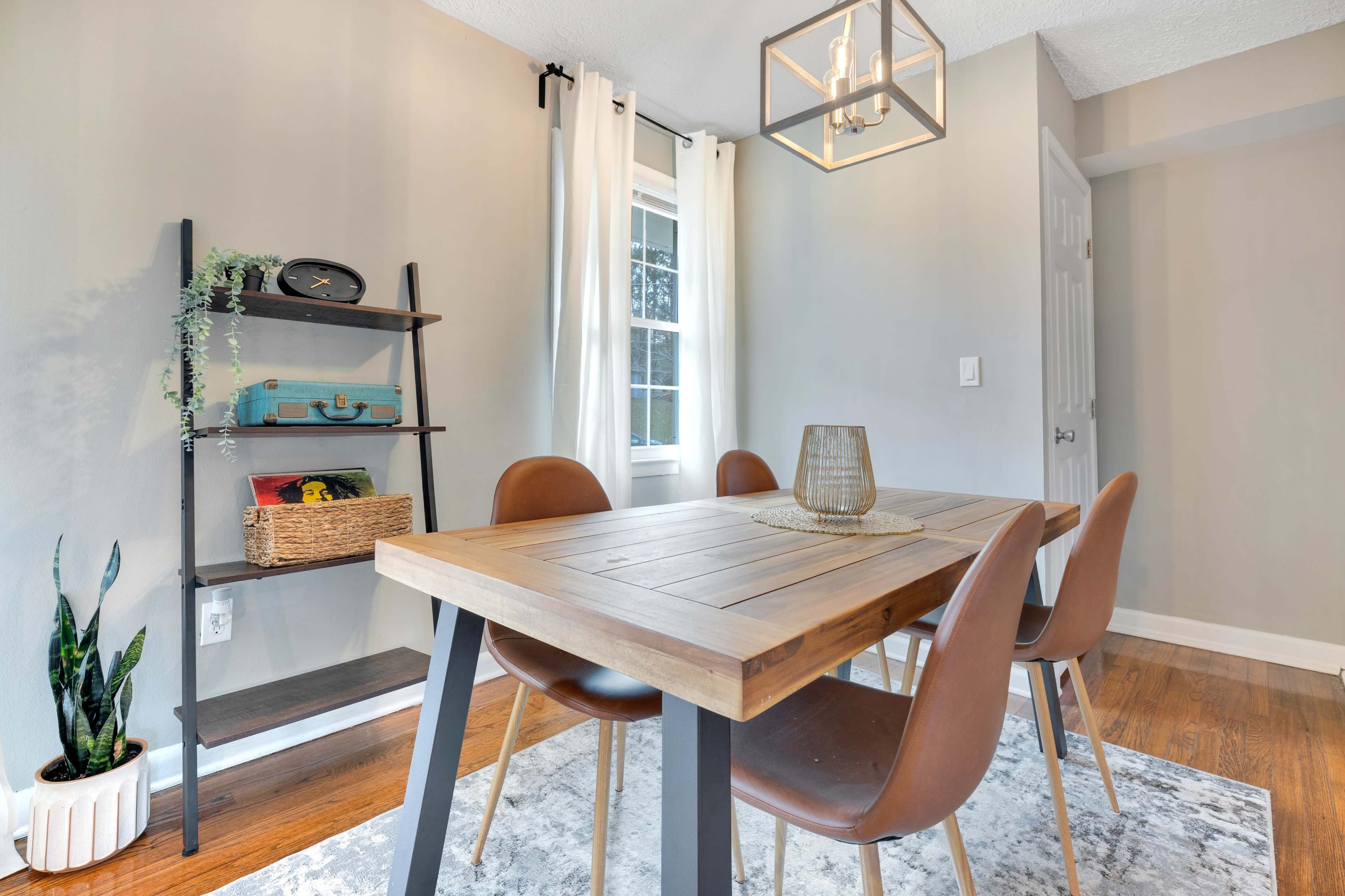 A dining room features a wooden table surrounded by brown chairs, a plant in a pot, and a shelving unit with decorative items.