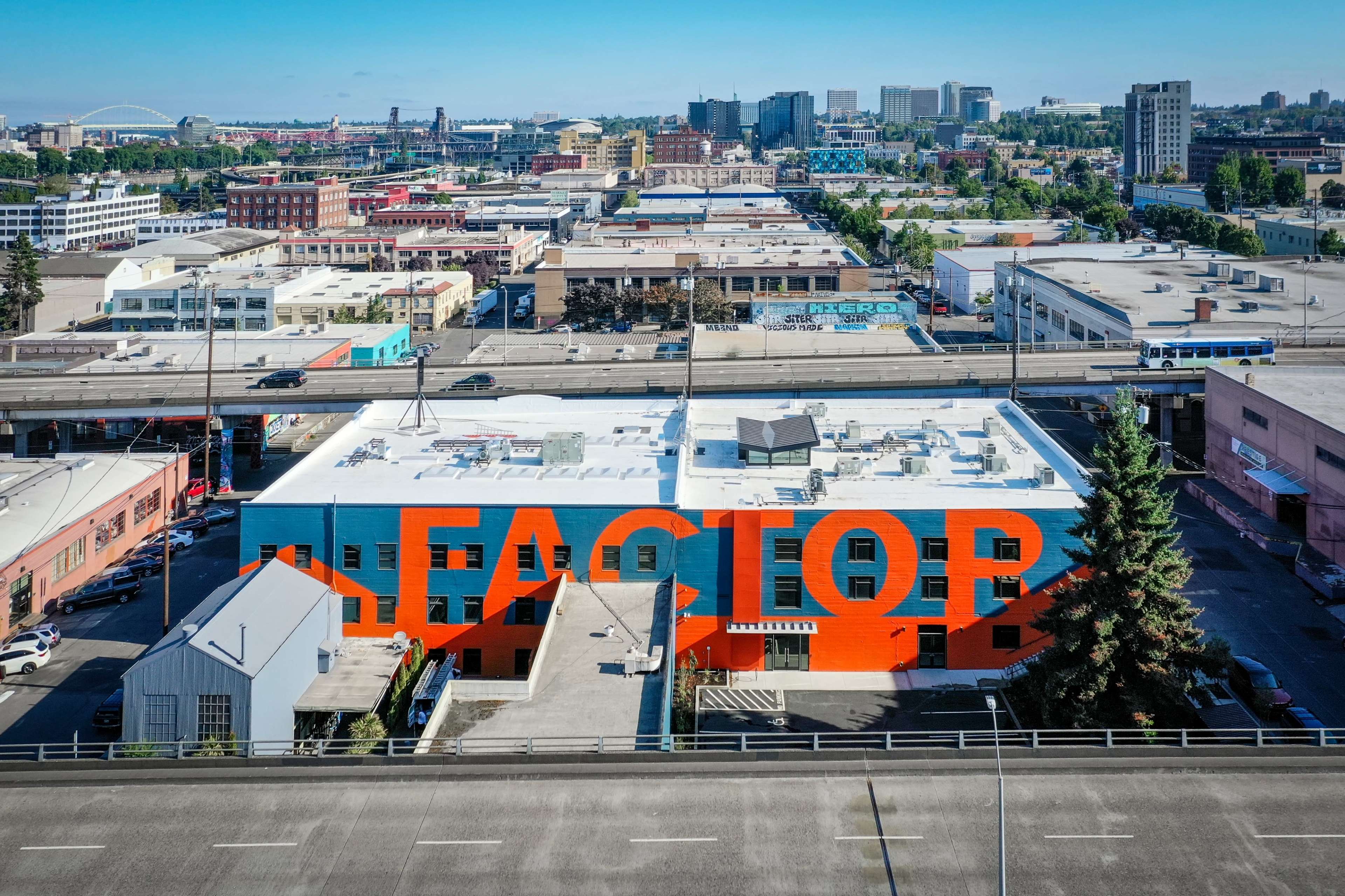 The image shows a vibrant orange and blue mural on a building with the word "FACTORY" prominently displayed, set against a backdrop of a city skyline and various structures.