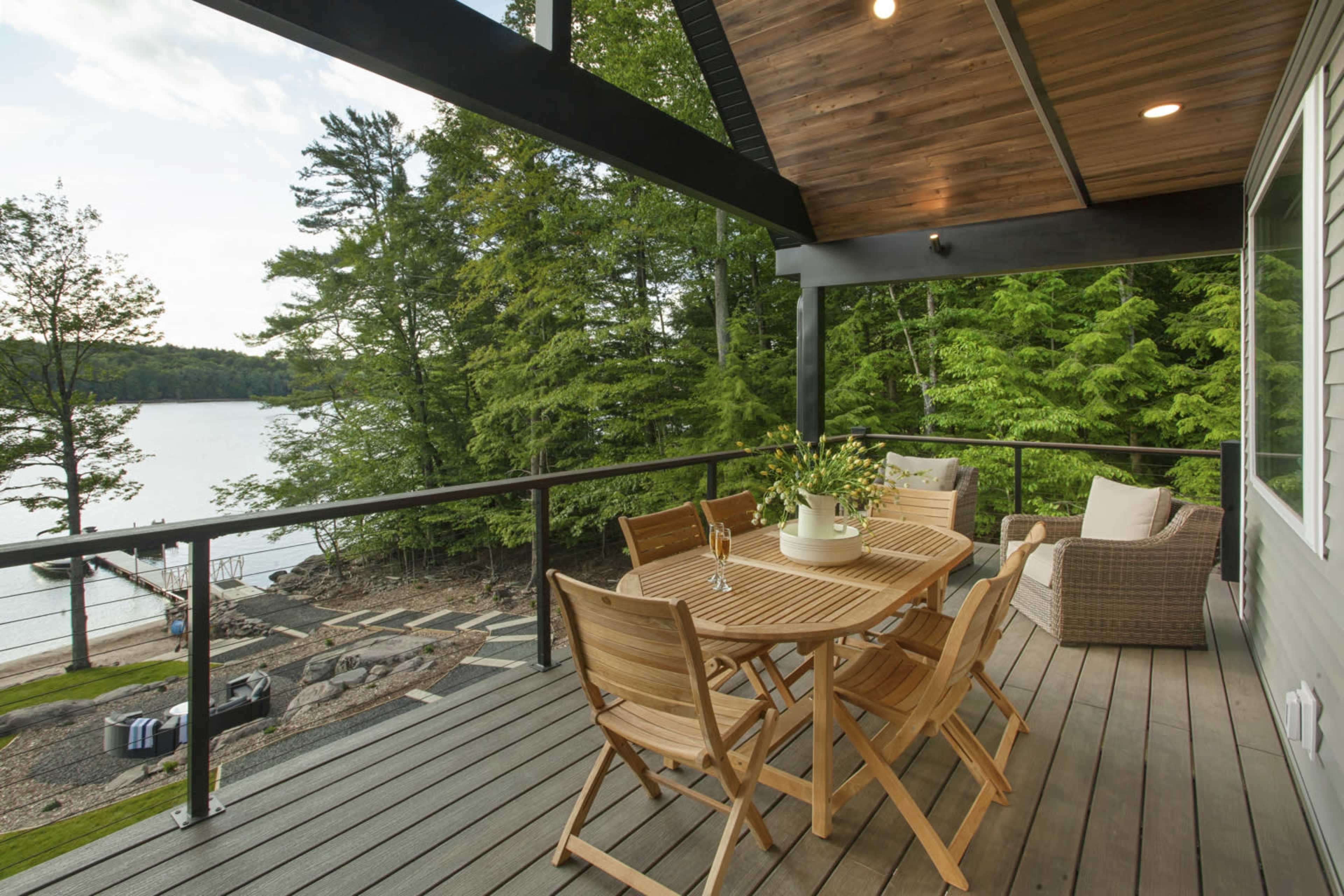 A wooden deck features a dining table and seating area, overlooking a calm lake surrounded by trees.