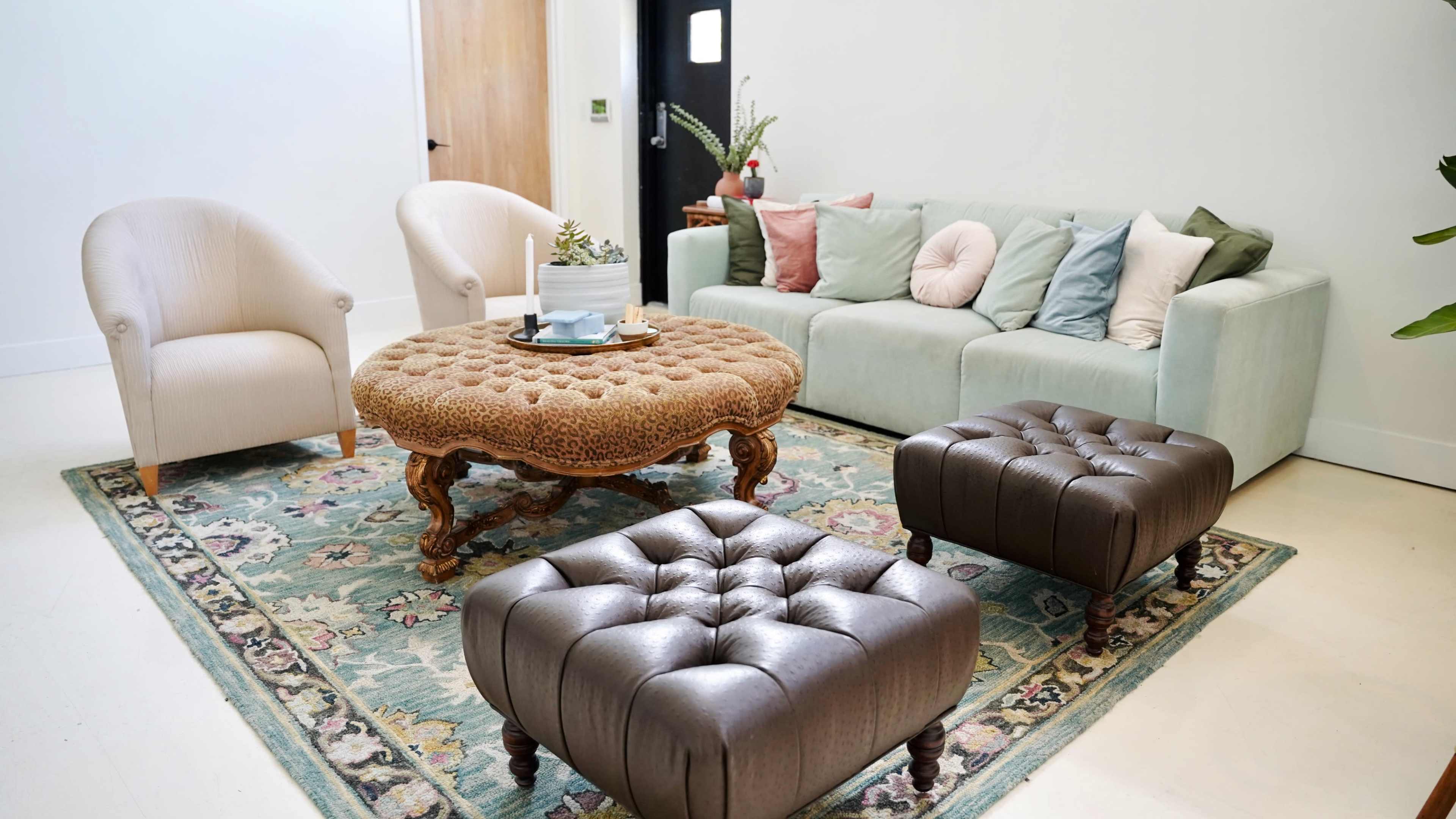 A living room features a light-colored sofa with assorted cushions, two tufted ottomans, and a round, textured coffee table, all arranged on a decorative rug.