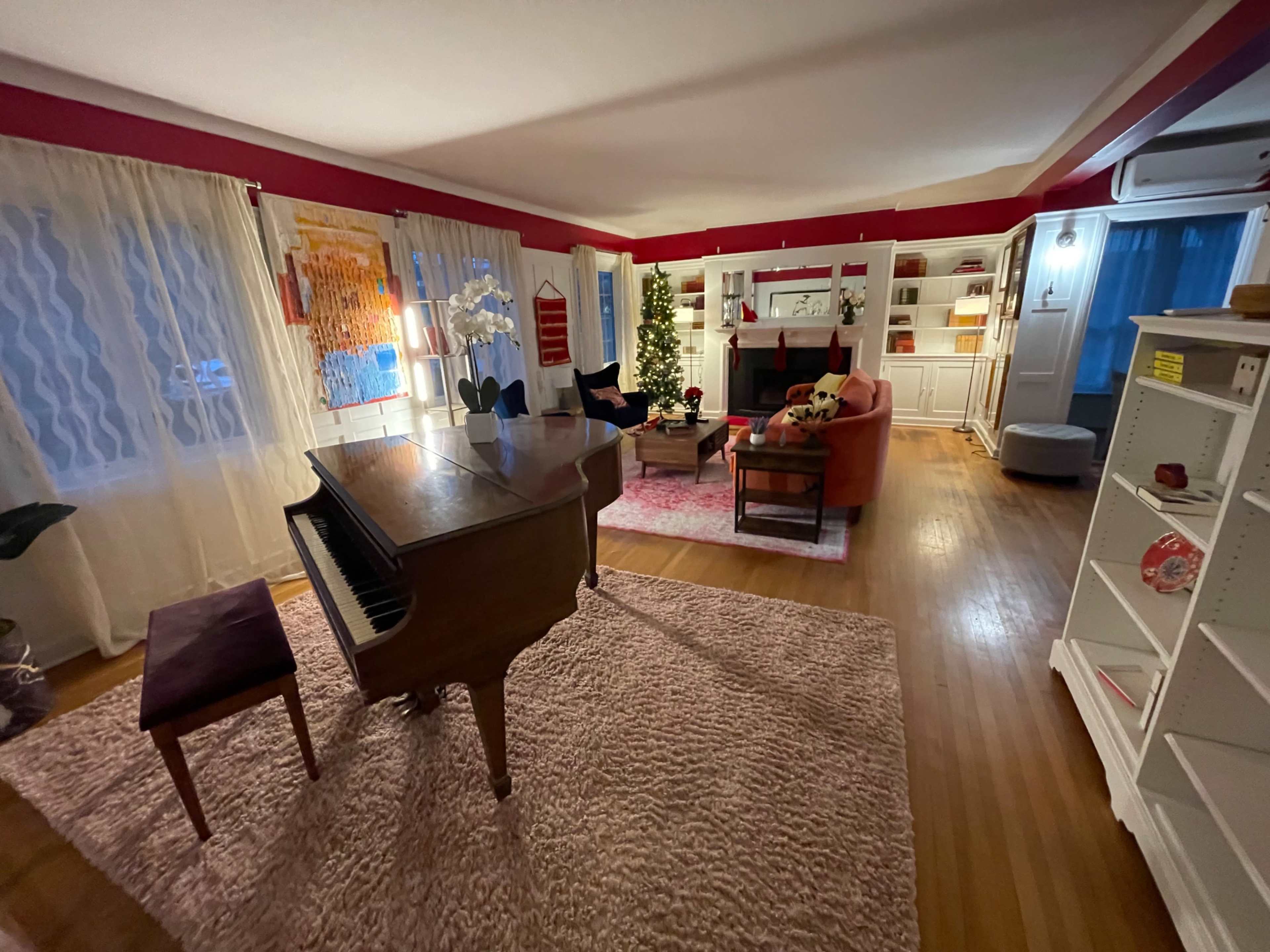 A spacious living room with a grand piano, a large area rug, and decorated shelves, featuring a Christmas tree and a cozy seating arrangement.