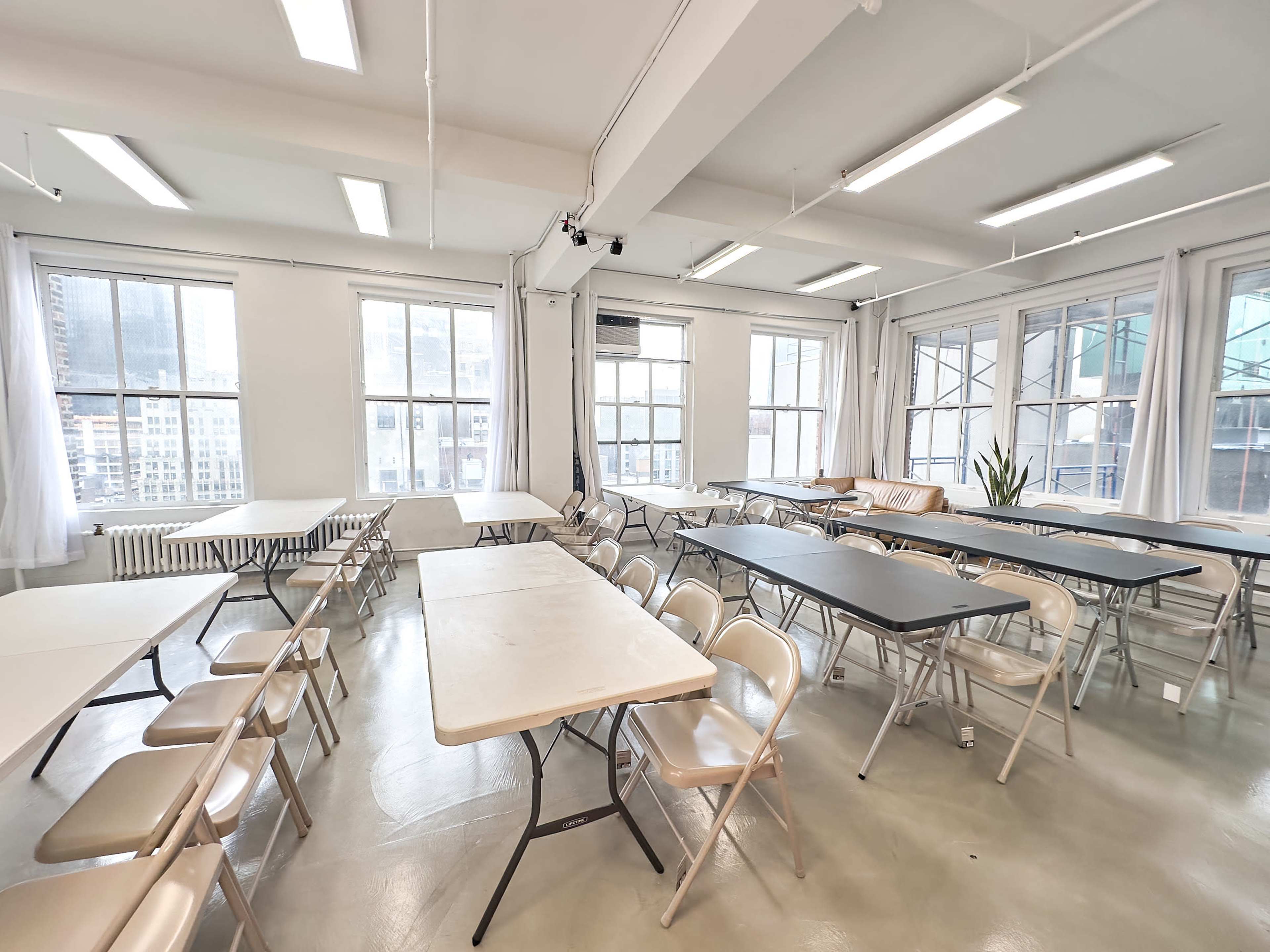 The image shows a spacious room with multiple folding tables and chairs arranged throughout, large windows allowing natural light, and minimal decorative elements.