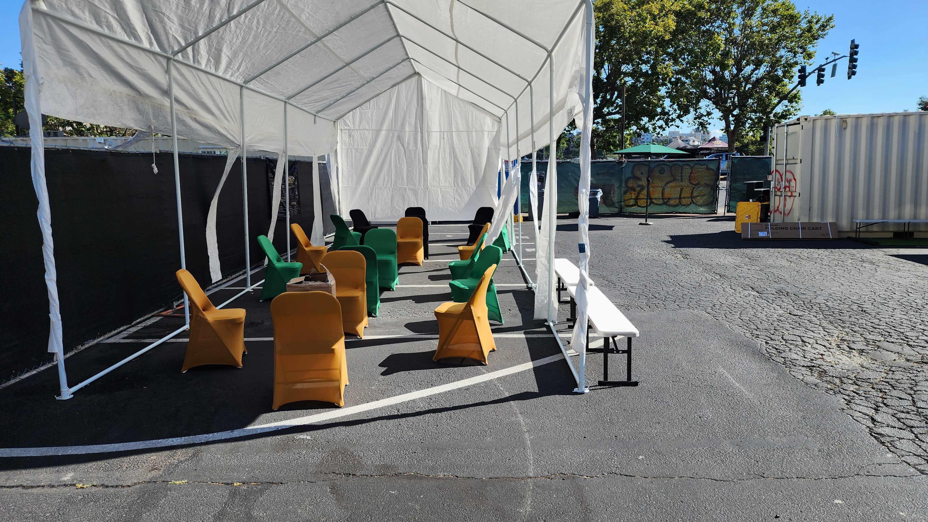 A canopy covers a row of tables and chairs set up on a paved outdoor area near a shipping container.