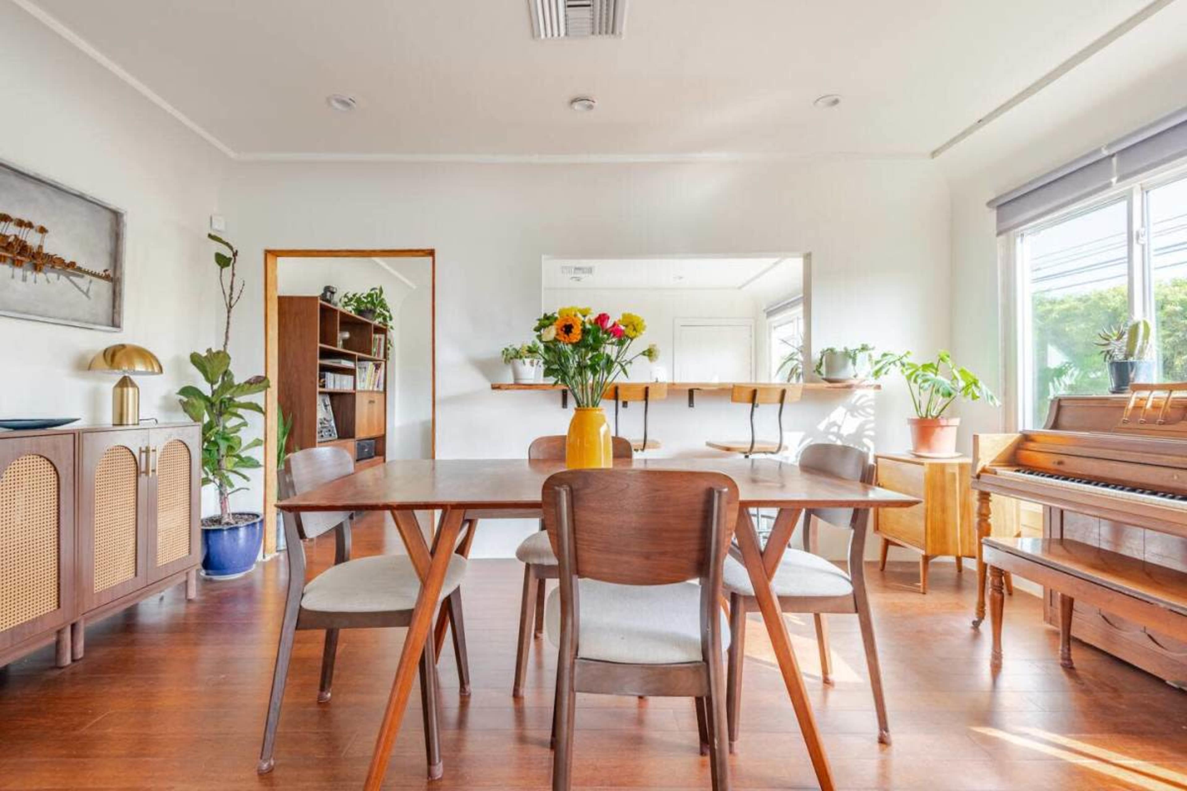 The image shows a bright dining area featuring a wooden table surrounded by four chairs, a vase of flowers on the table, and a piano in the corner along with houseplants and a sideboard.