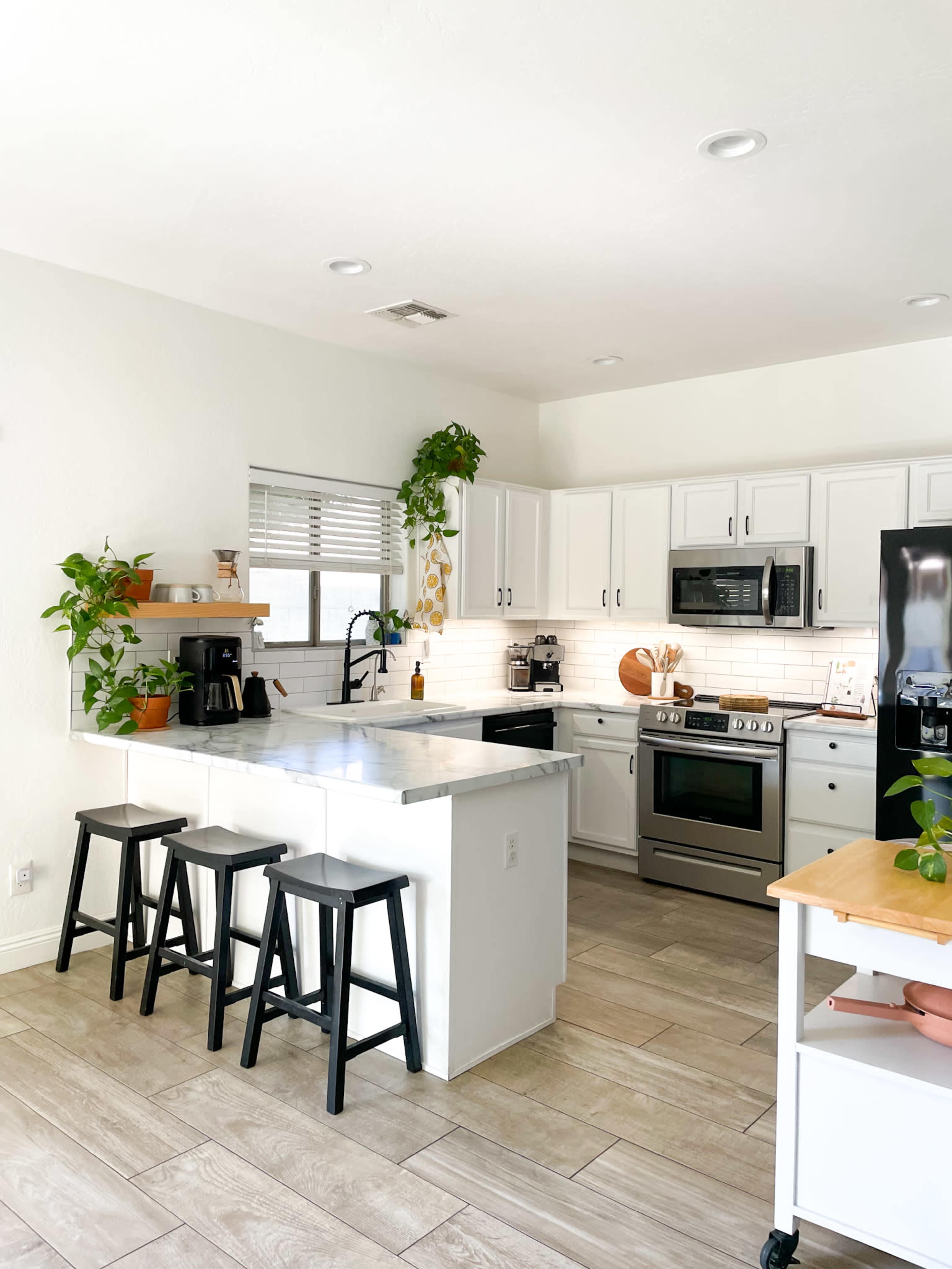 The kitchen features white cabinetry, a marble countertop island with three black stools, and several indoor plants.