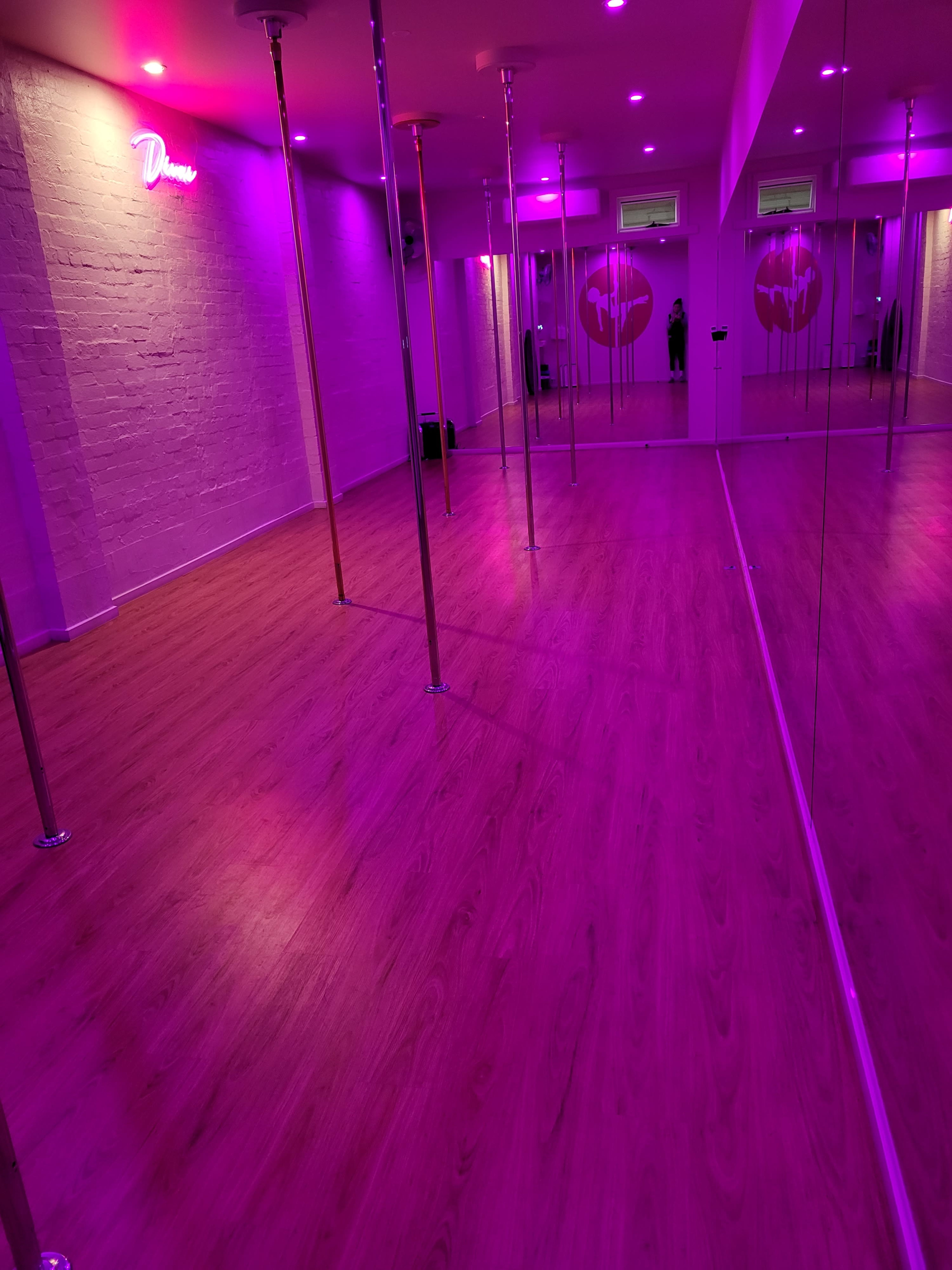 A brightly lit dance studio with pink neon lights, wooden flooring, and several mirrors reflecting tall poles.