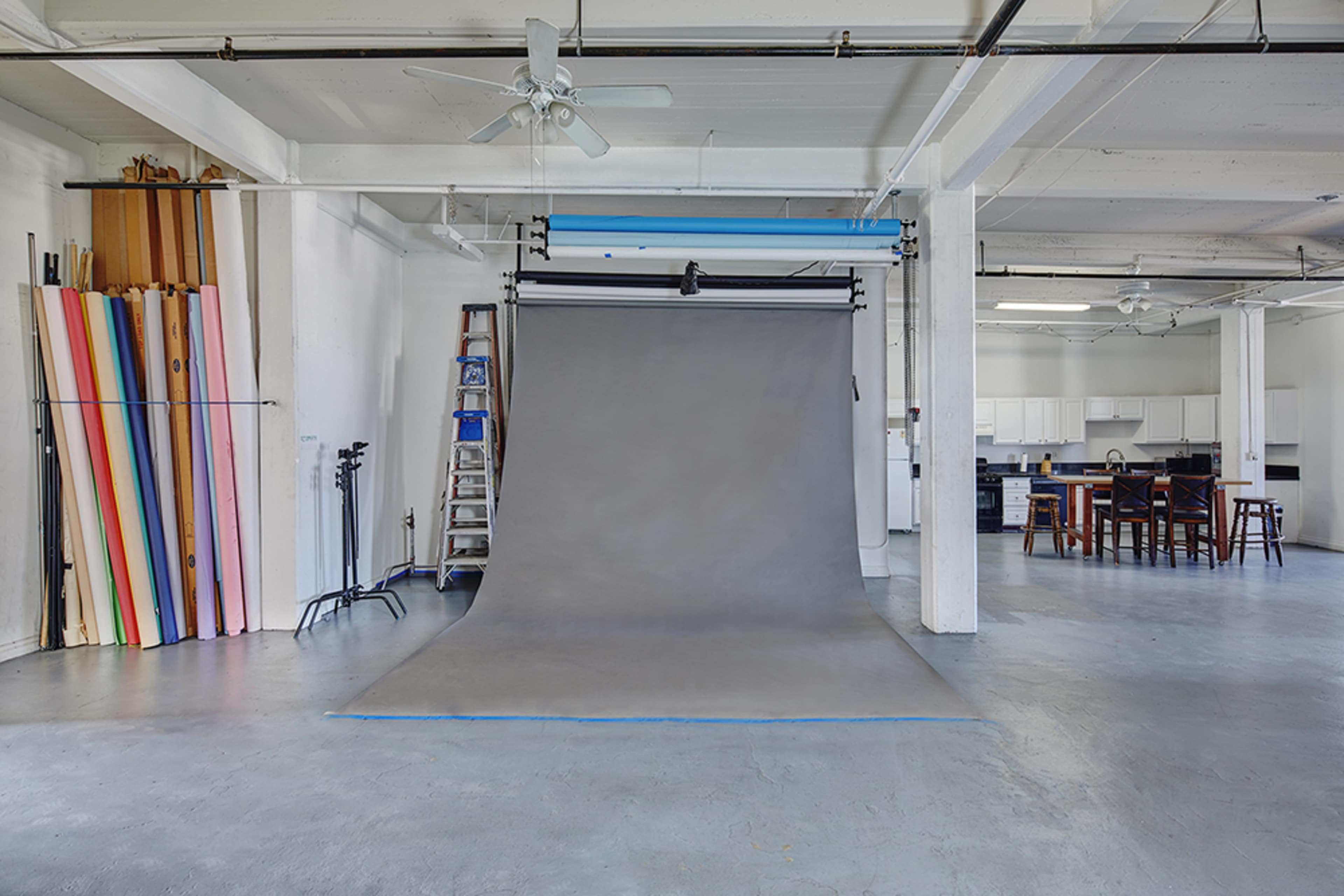 A spacious photography studio features a gray backdrop, a tall ladder, colorful backdrop rolls, and a kitchenette visible in the background.