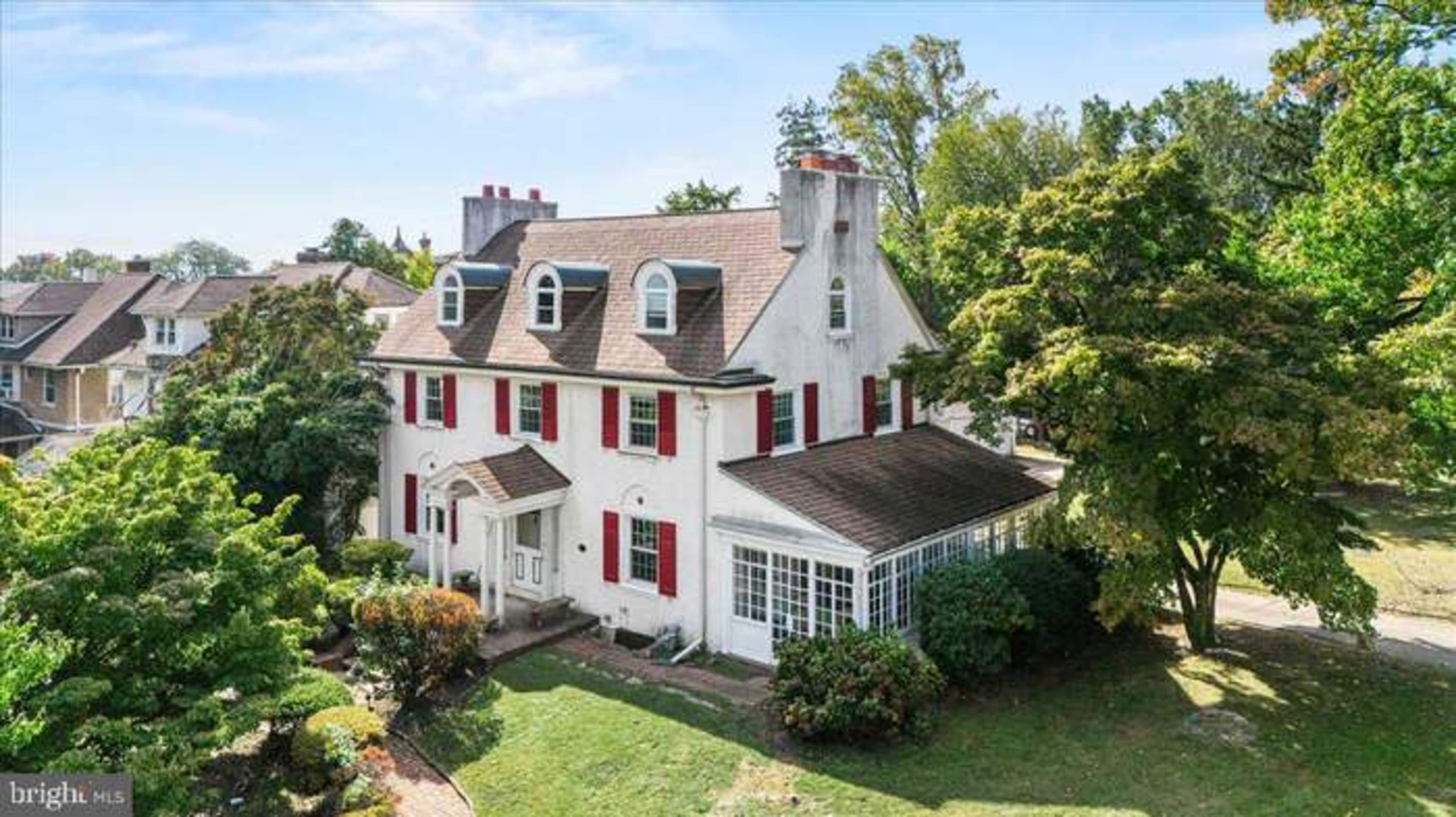 A two-story white house with a pitched roof and red shutters is surrounded by trees and shrubs.