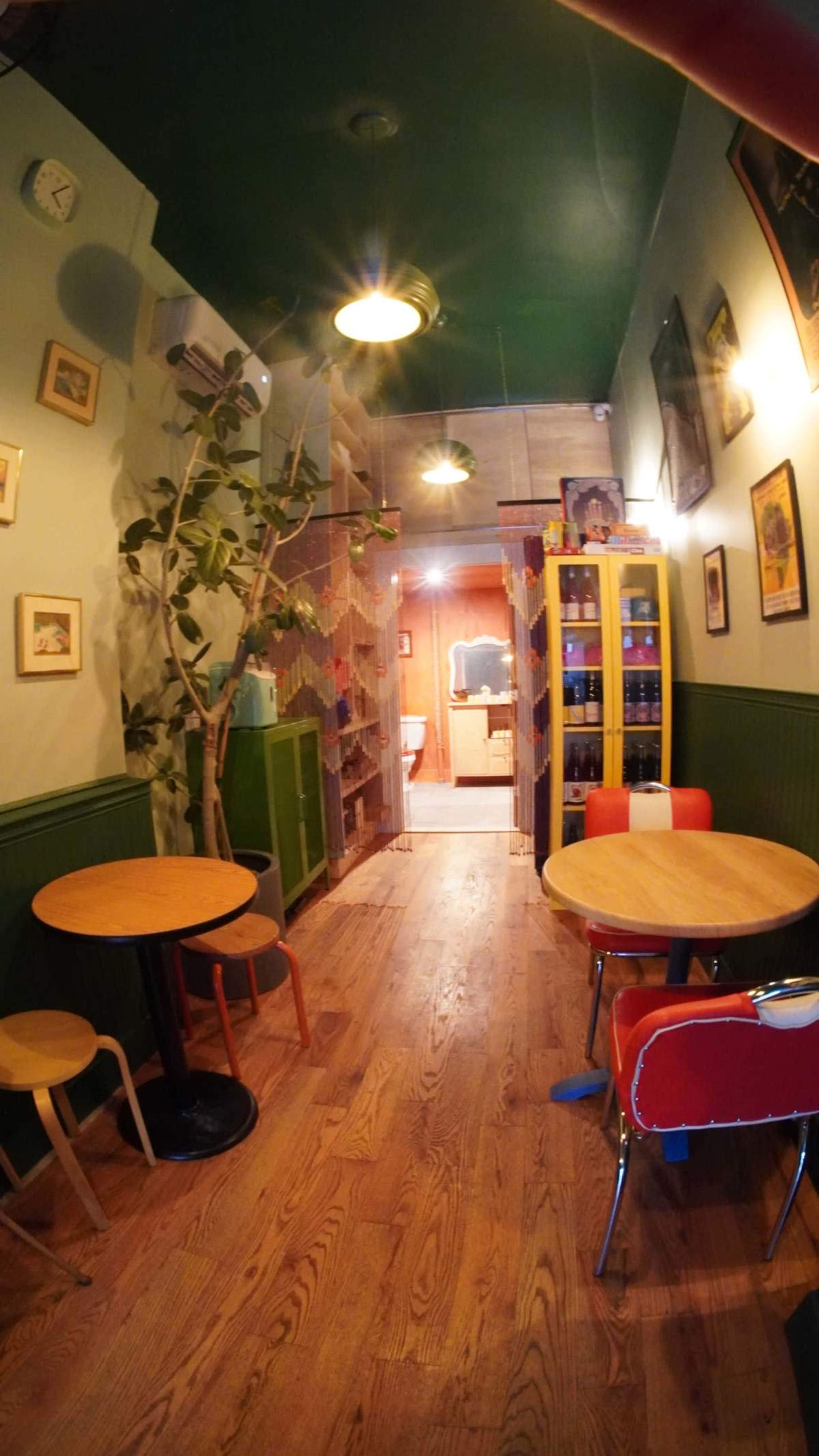 The image shows a narrow café interior with wooden flooring, two small tables, and a colorful partition leading to a restroom area.