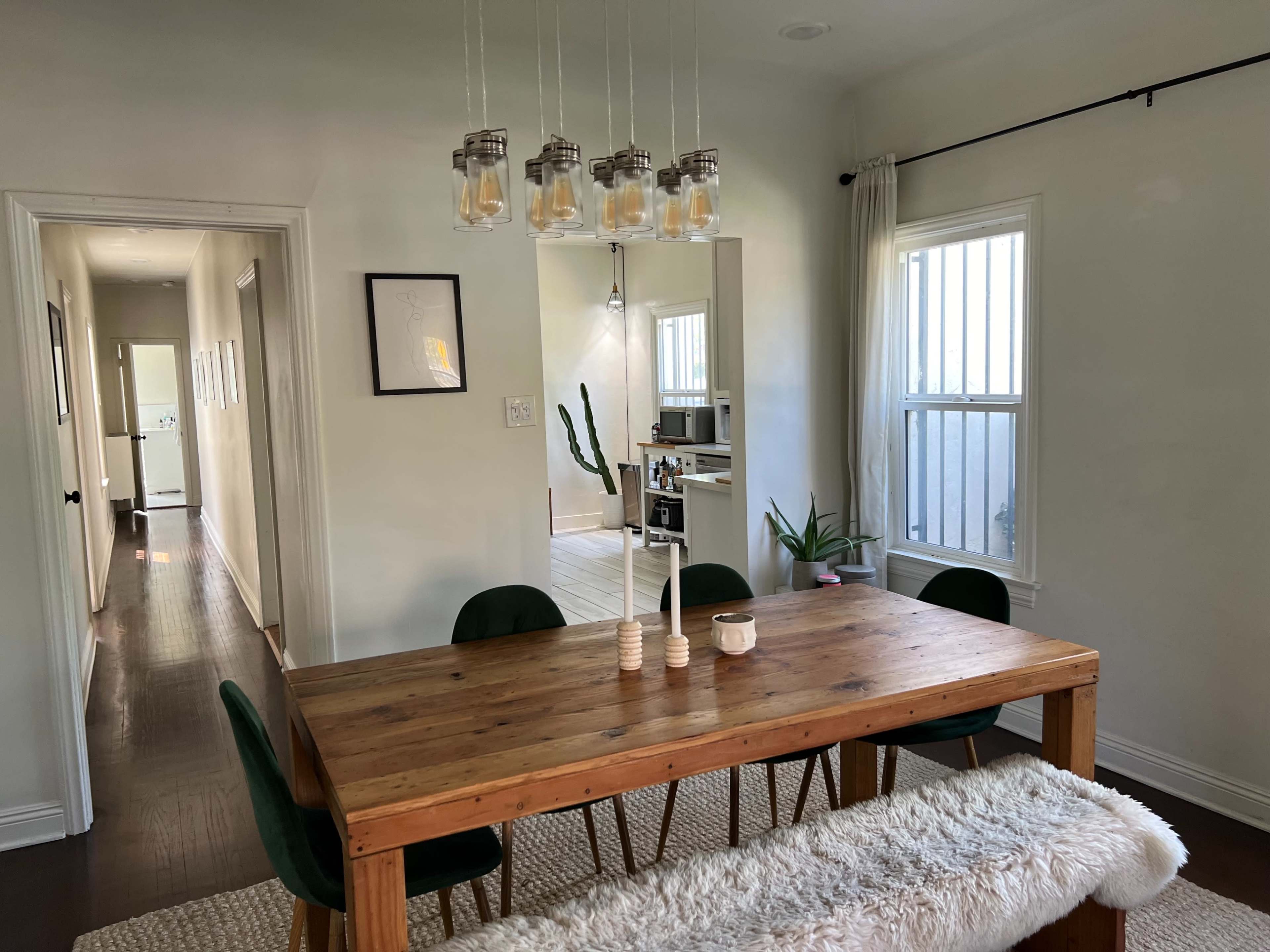 A wooden dining table with green chairs is set under a hanging light fixture in a well-lit room, with a hallway and kitchen visible in the background.
