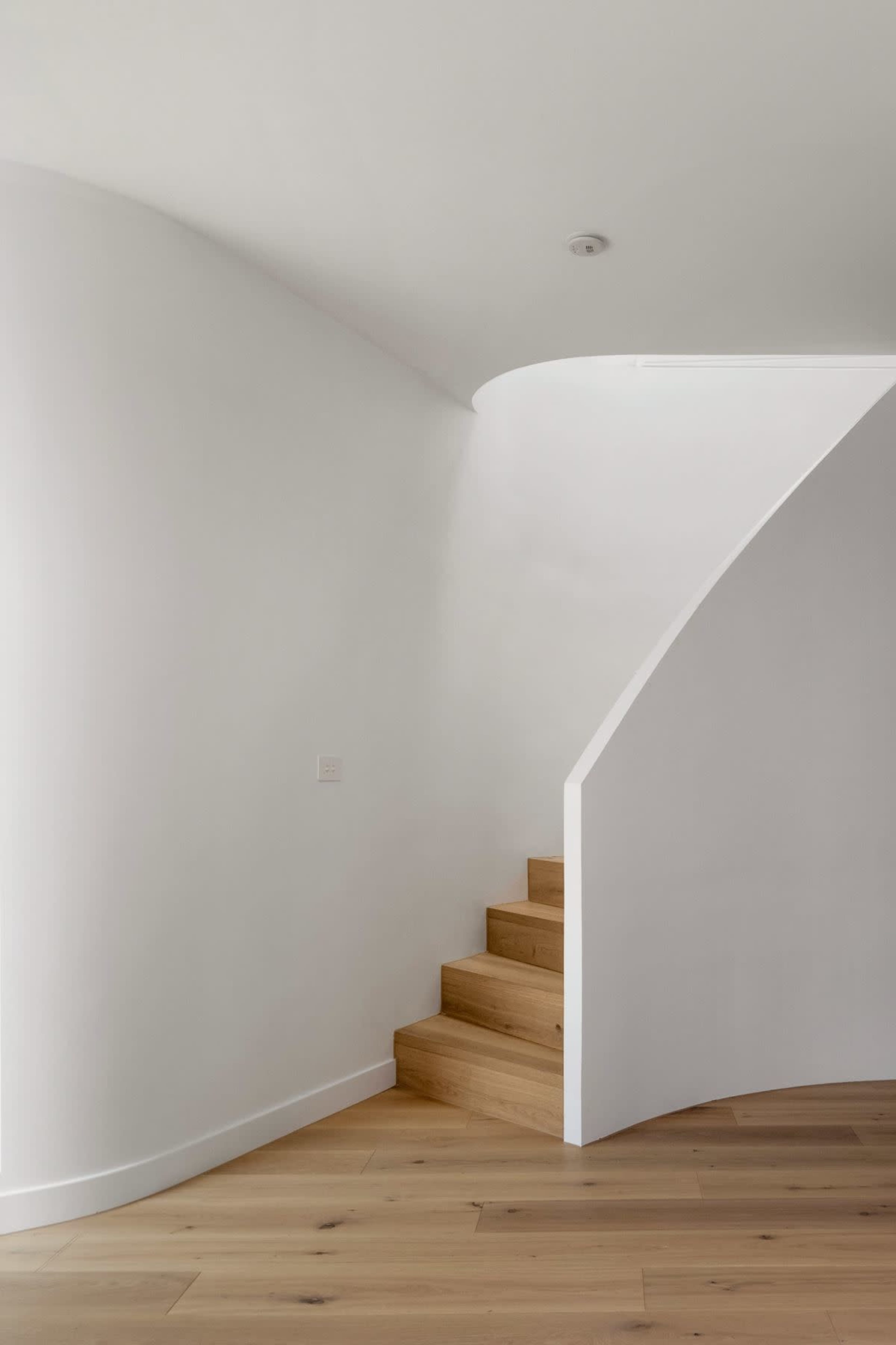 A curved staircase with wooden steps leads upward, situated against a smooth white wall in a minimalist interior space.