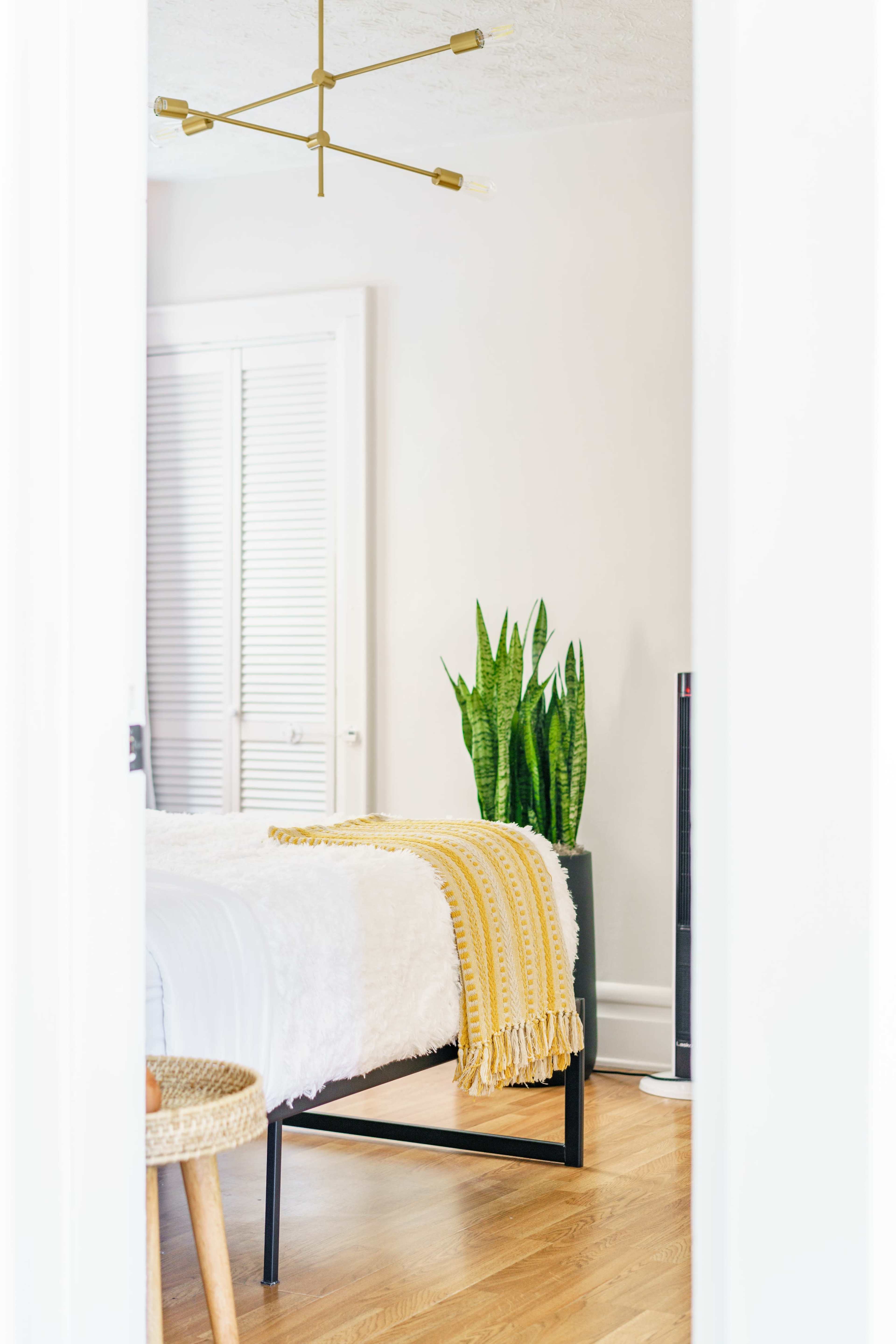 A cozy bedroom features a bed with a white coverlet, a yellow-striped throw, a potted plant, and a minimalist light fixture.