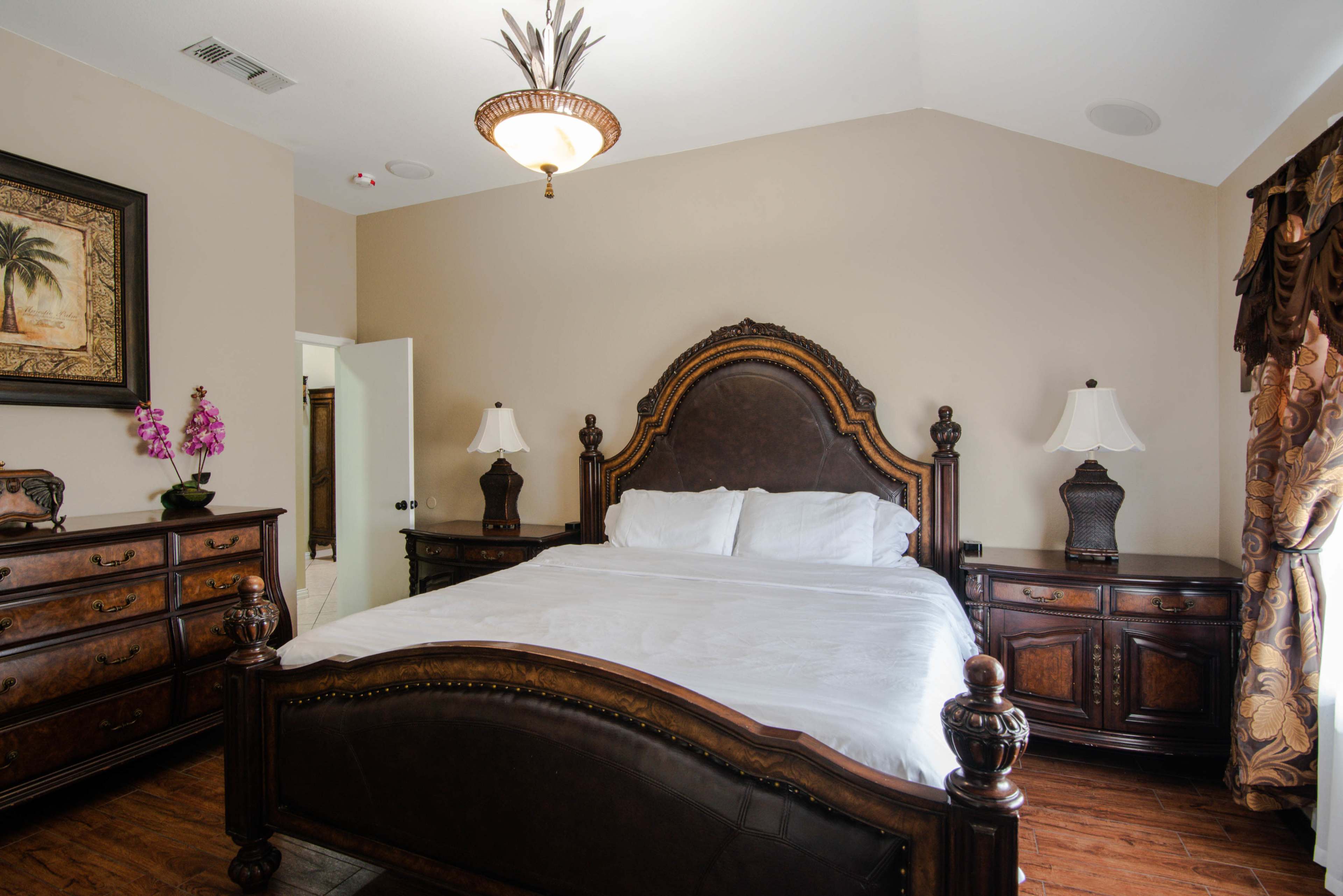 A spacious bedroom features a large wooden bed with a dark headboard, flanked by two bedside lamps, and decorated with a painting and flowers.