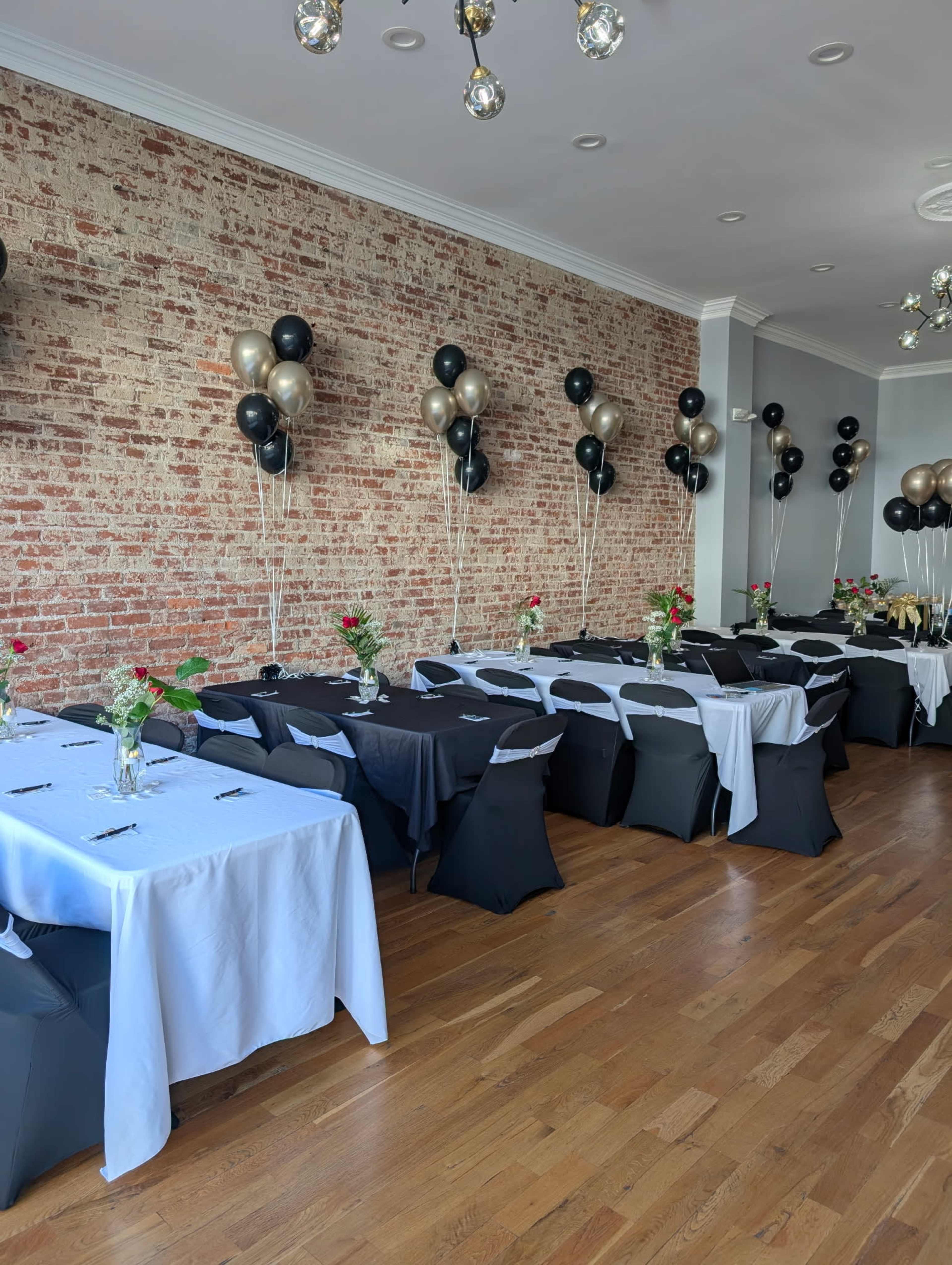 A spacious event room features rows of tables dressed in black and white linens, adorned with vases of flowers and black, gold, and silver balloons hovering above.