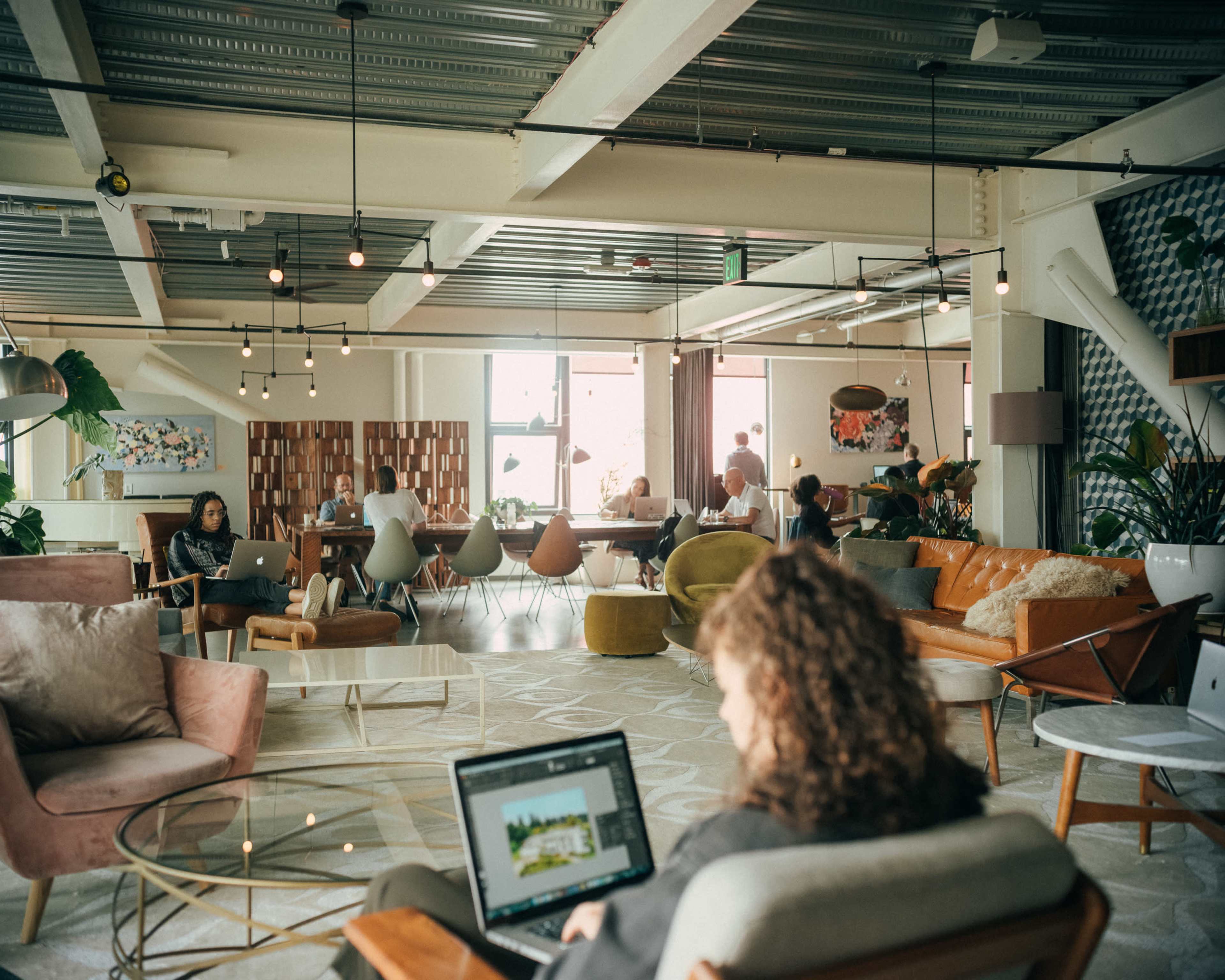 A modern coworking space features people working on laptops amidst various seating arrangements and plants.
