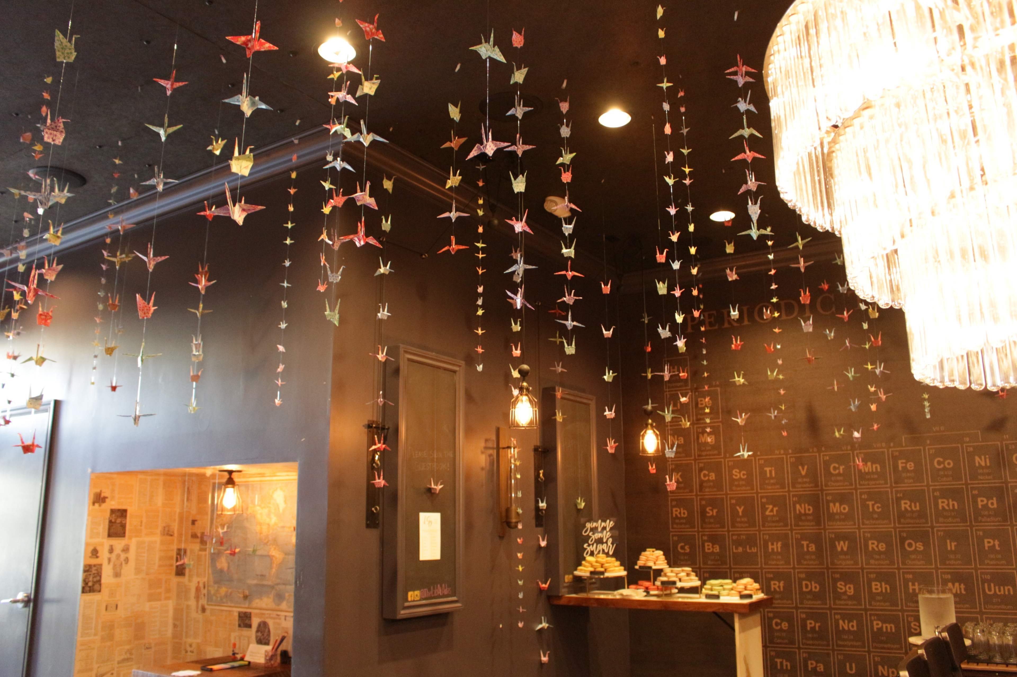 The interior of a café features many hanging paper cranes and a large chandelier above a counter with pastries.