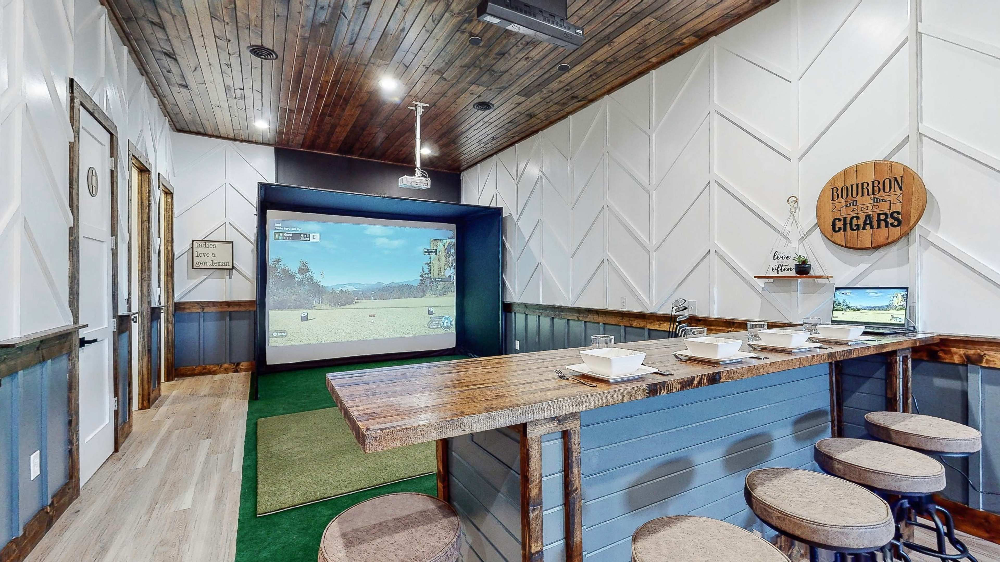 A home entertainment room featuring a projection screen with a golf simulation, a wooden bar with stools, and a green carpeted area.