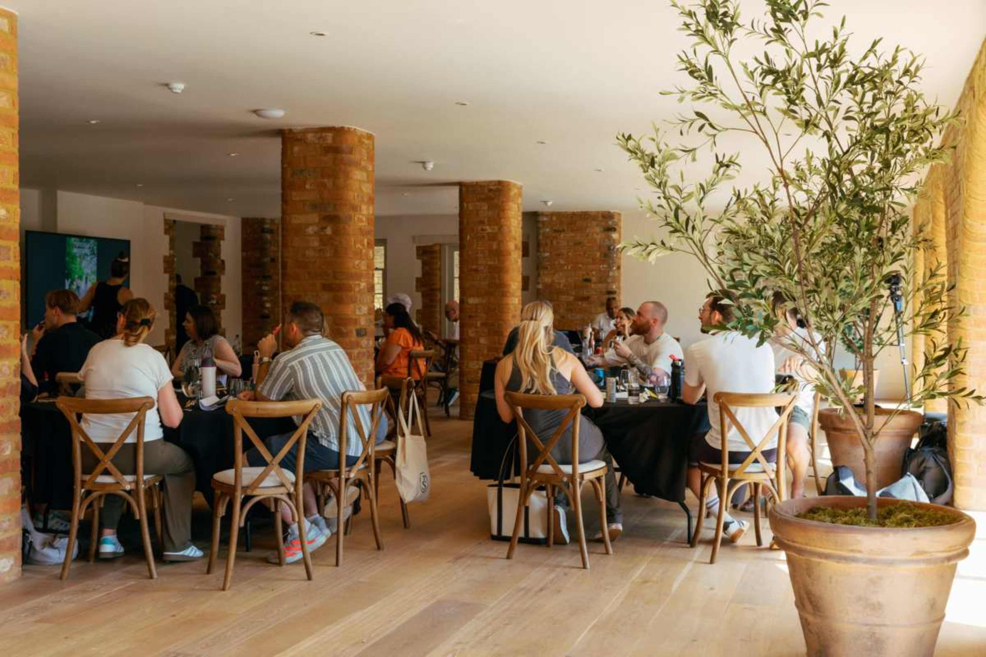A group of people sits at tables in a spacious dining area with brick pillars and natural light.