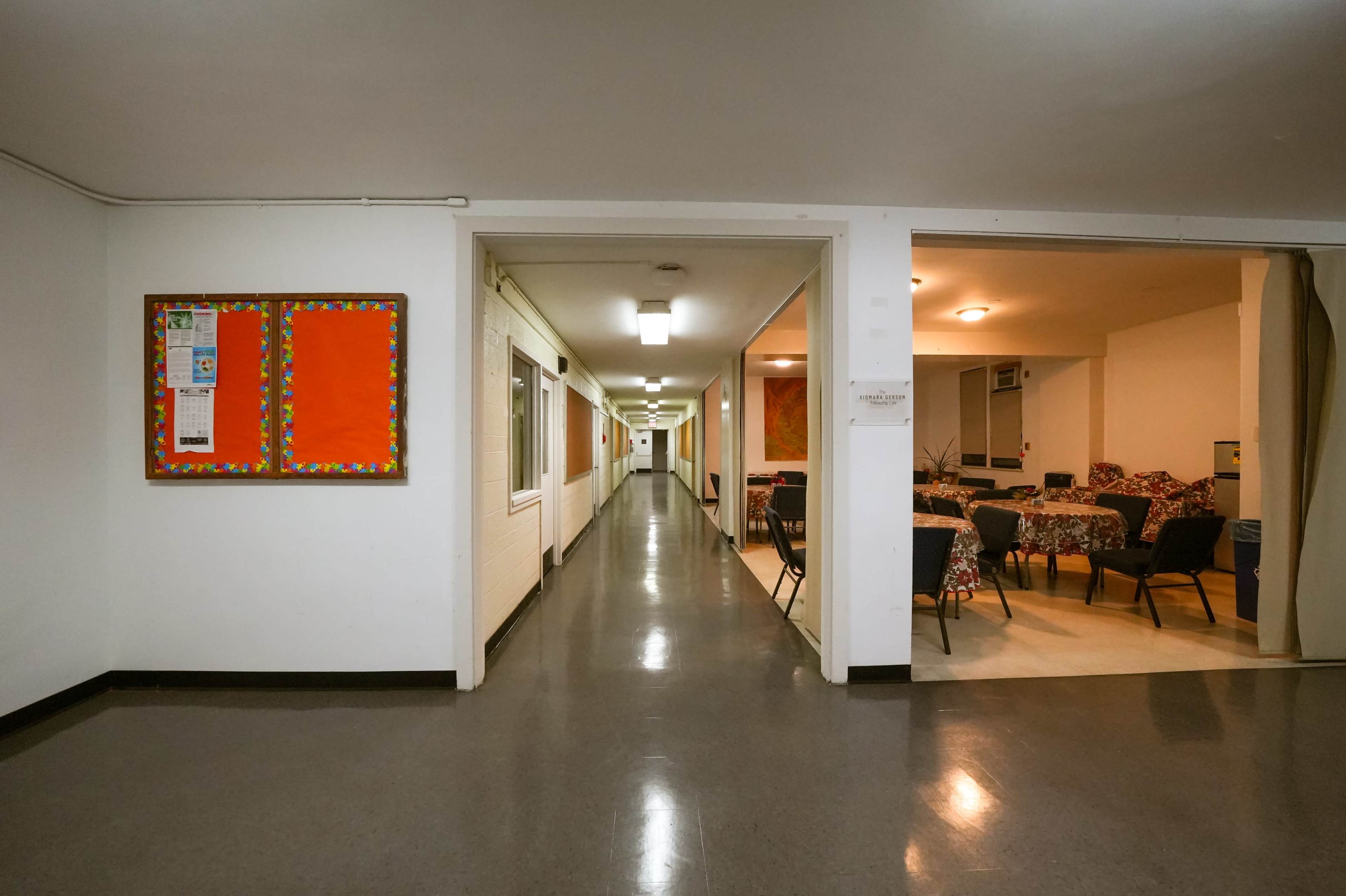 A long hallway leading to a room with tables and chairs, with bulletin boards on the wall to the left.