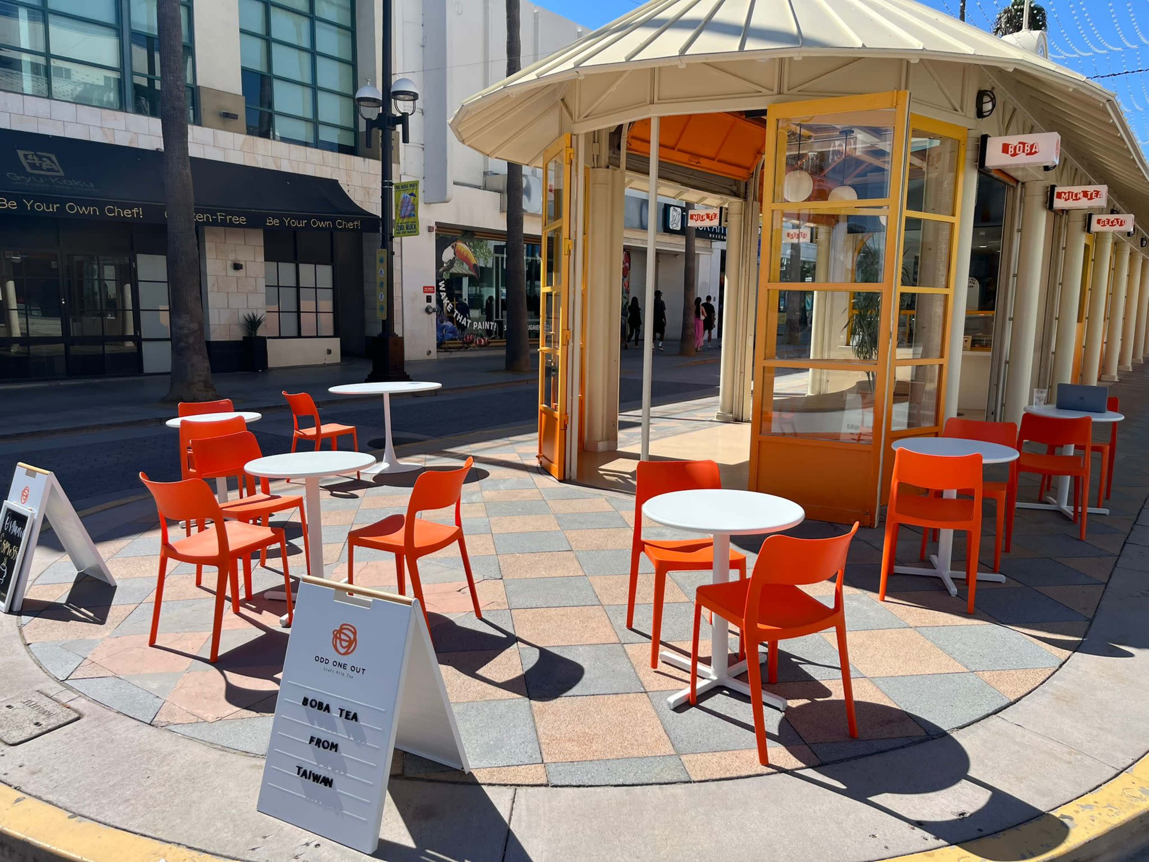 The image shows an outdoor seating area with orange chairs and white tables, located in a circular structure with large windows.