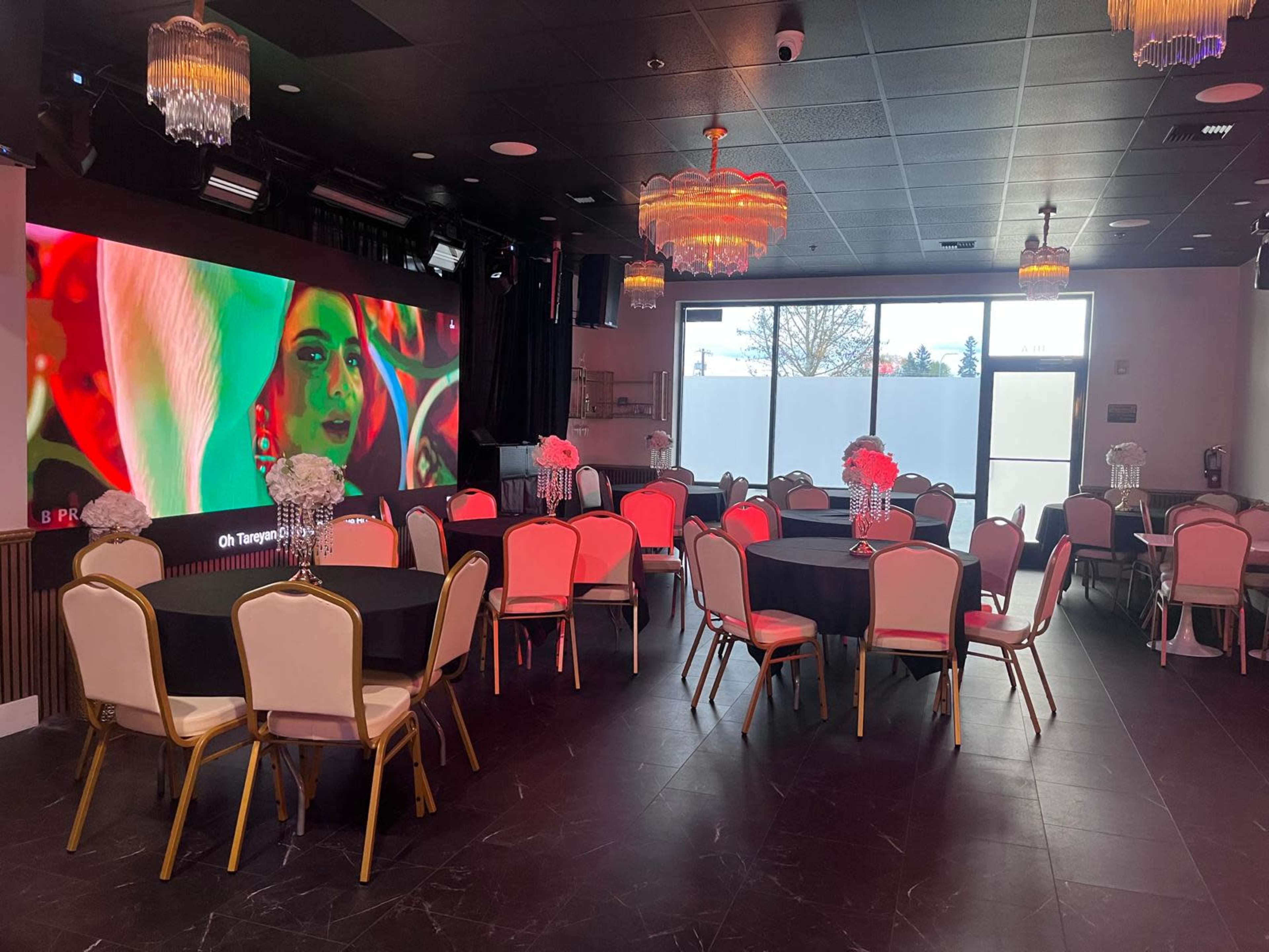 The image shows a modern event space with round tables set for dining, adorned with floral centerpieces, and a large screen displaying vibrant visuals in the background.