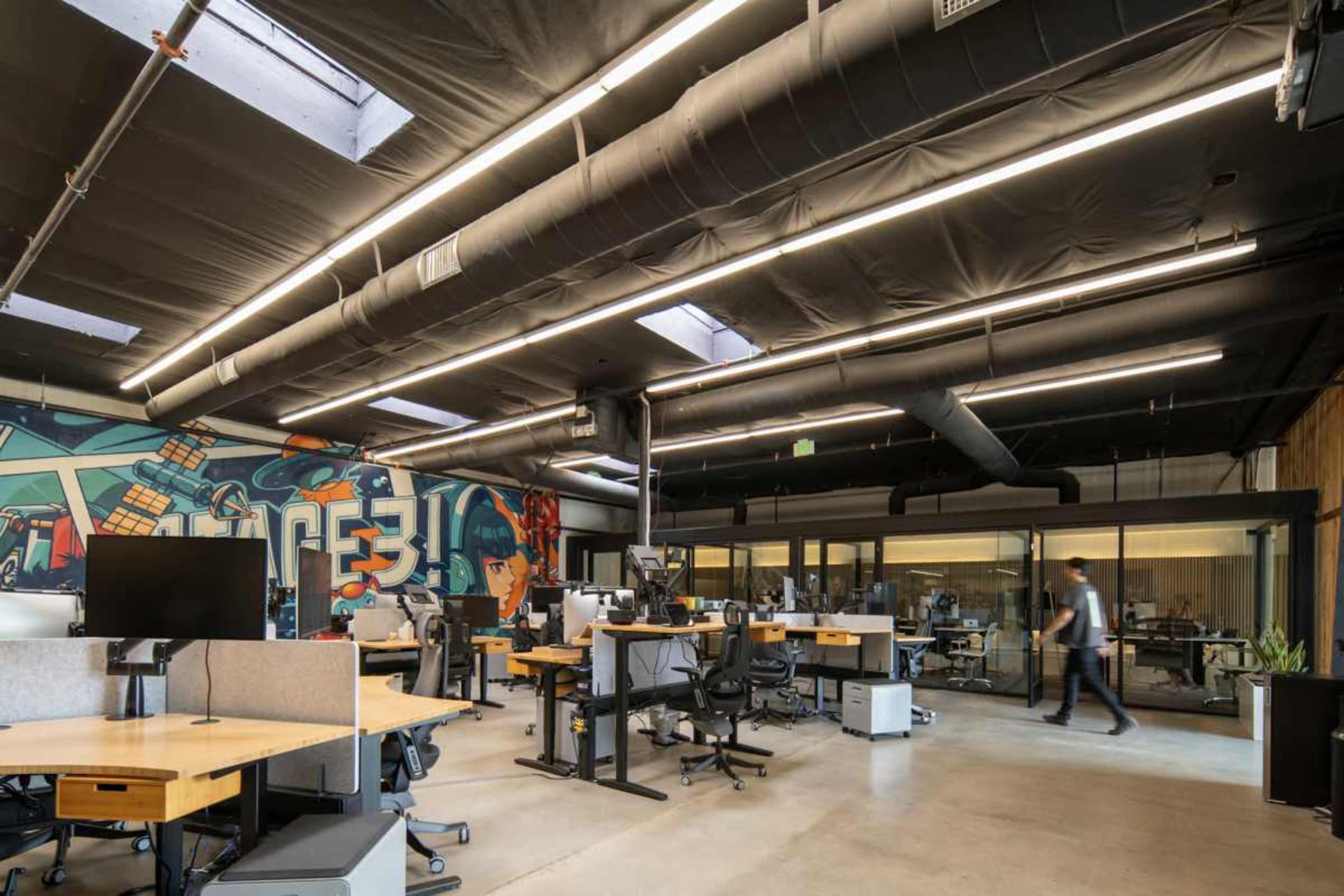 The image shows a modern office space with a mural on one wall, featuring several desks and a few people working.