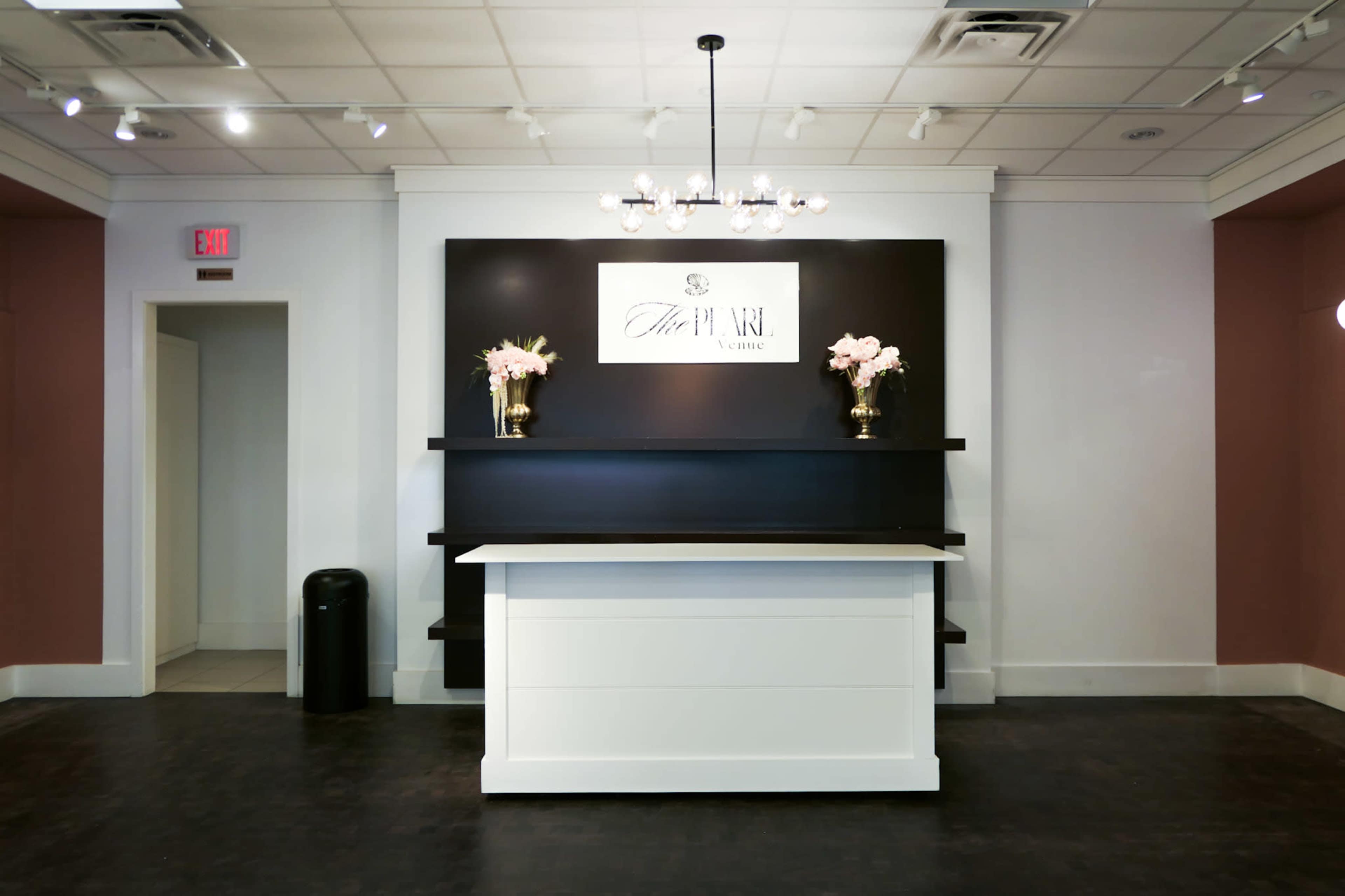 The image features a modern reception area with a white front desk against a black wall displaying a logo, flanked by decorative vases with flowers.