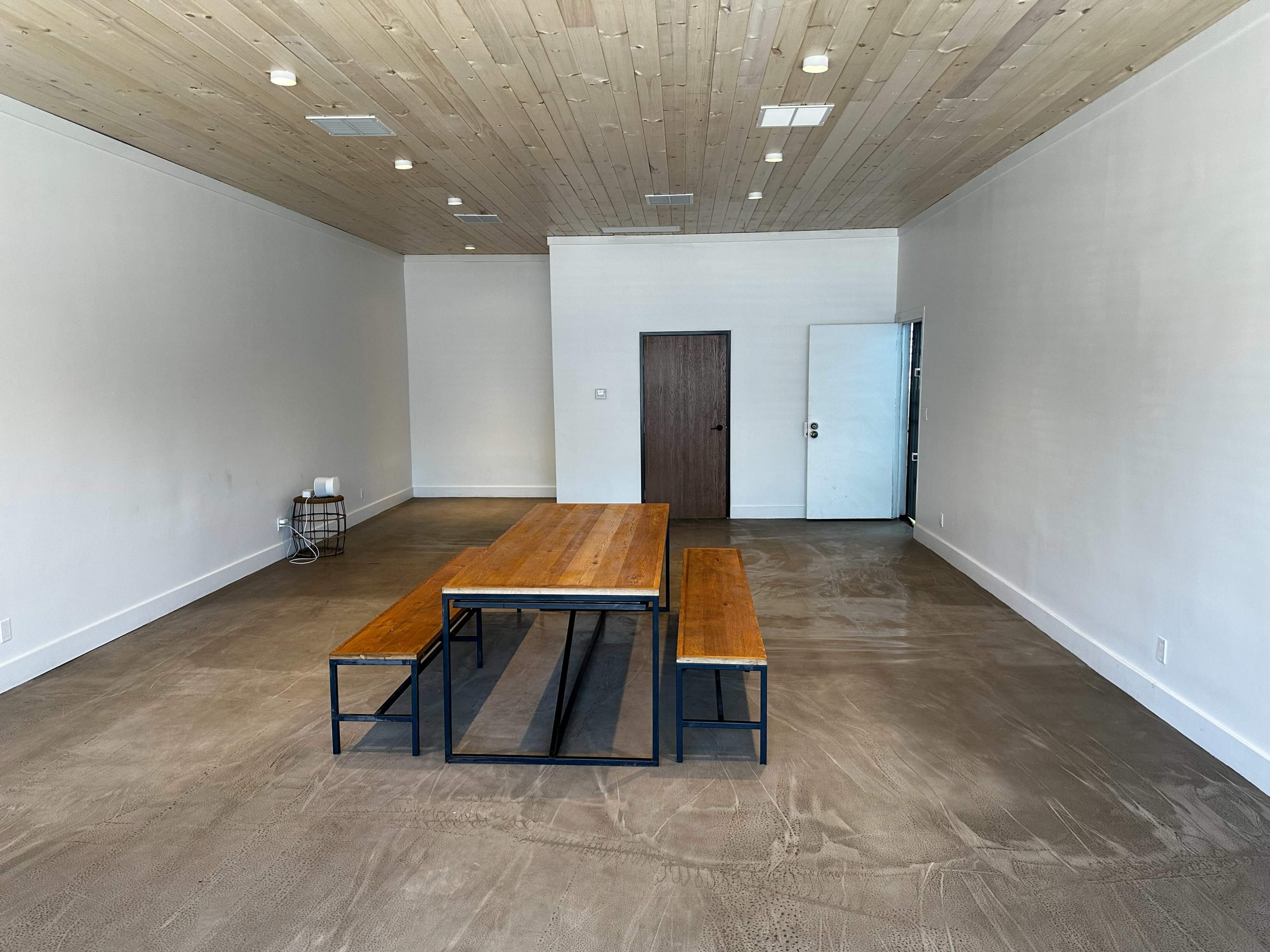 A spacious, minimalistic room featuring a large wooden table with metal legs, concrete flooring, and a single door on the back wall.