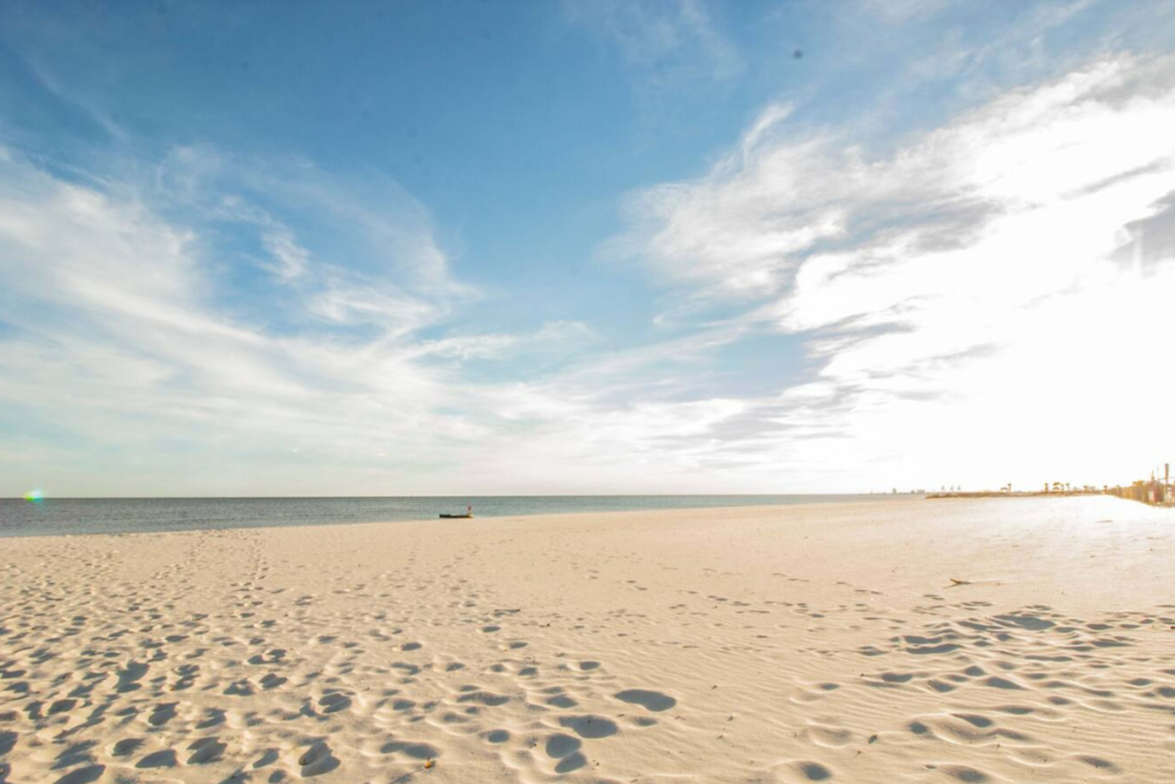 A sandy beach stretches out under a blue sky, with gentle waves lapping at the shore and distant buildings lining the horizon.