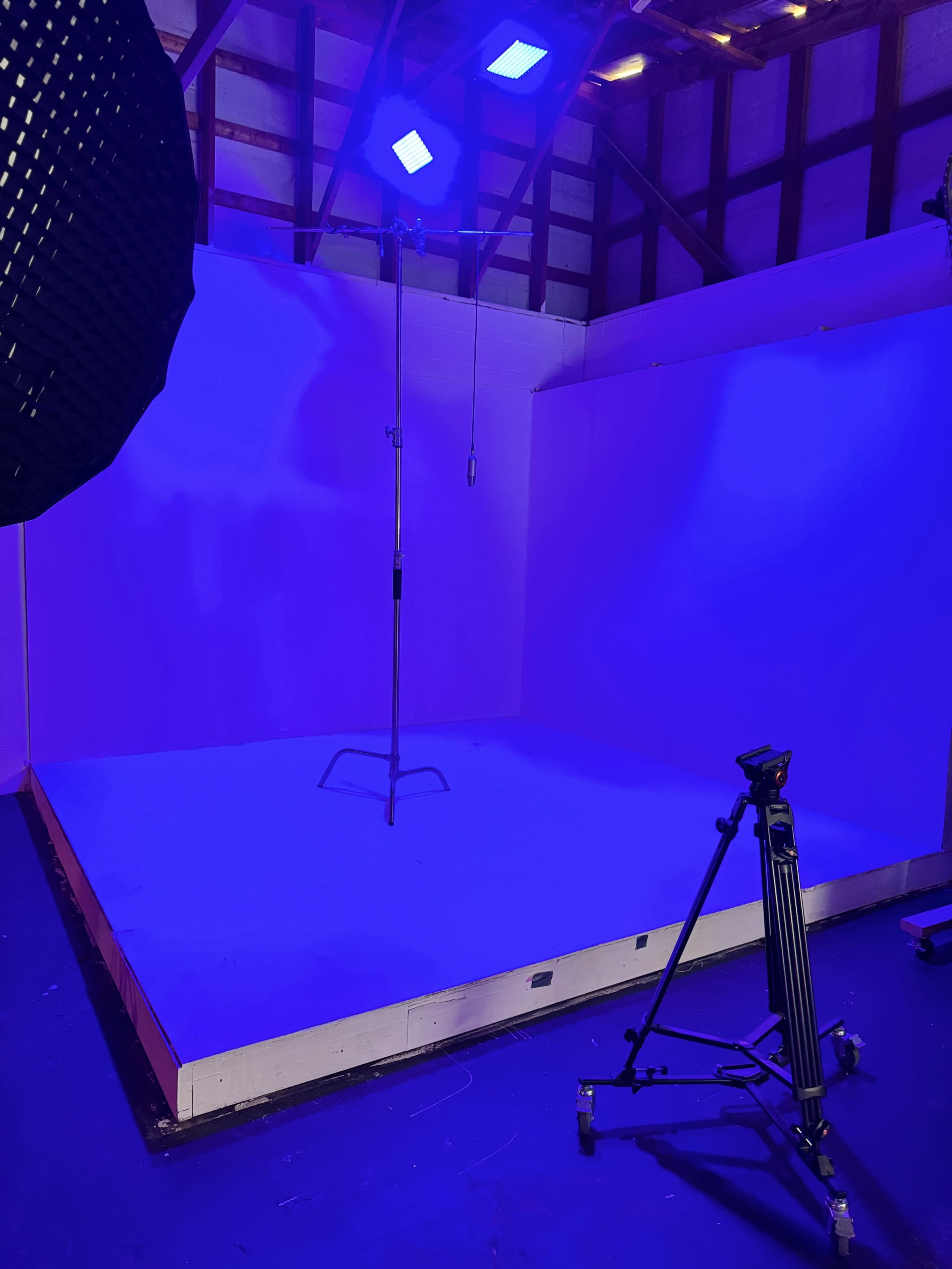 The image shows an empty studio space with a seamless backdrop, a lighting setup, and a stand in a dimly lit environment illuminated by blue lights.