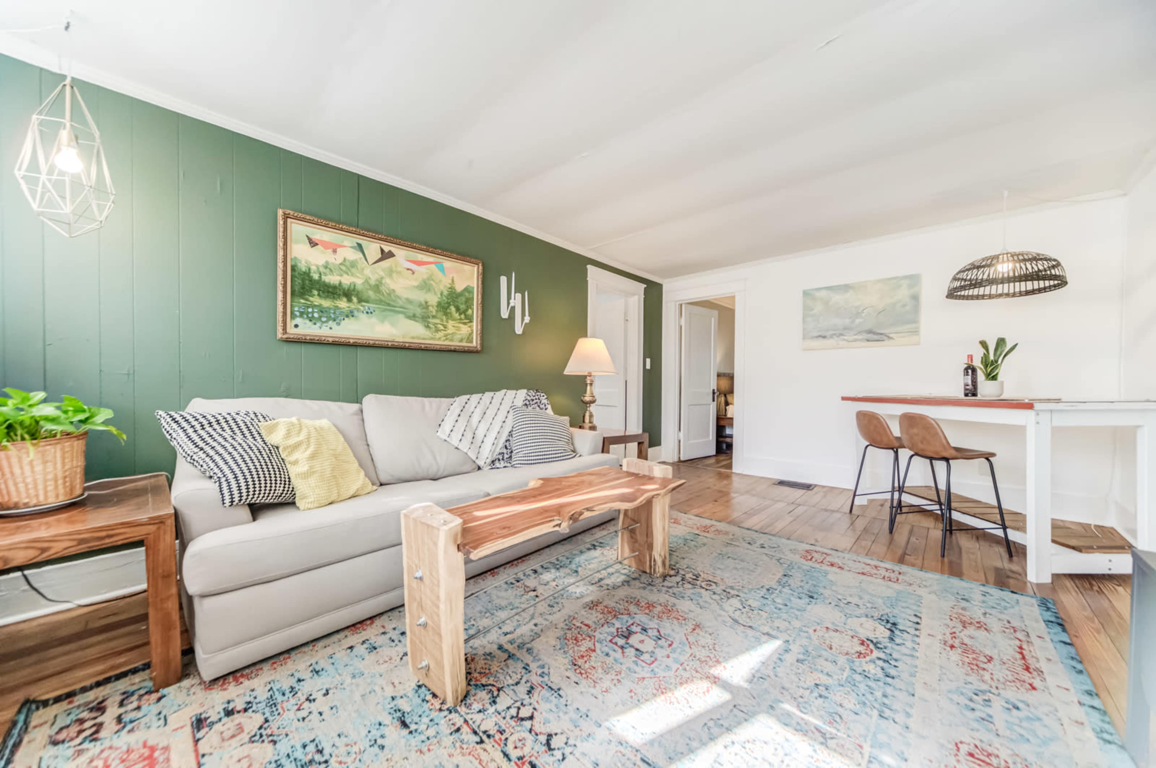 A living room featuring a light gray sofa with decorative pillows, a wooden coffee table, and a patterned rug, all arranged around a wall painted in green with artwork.