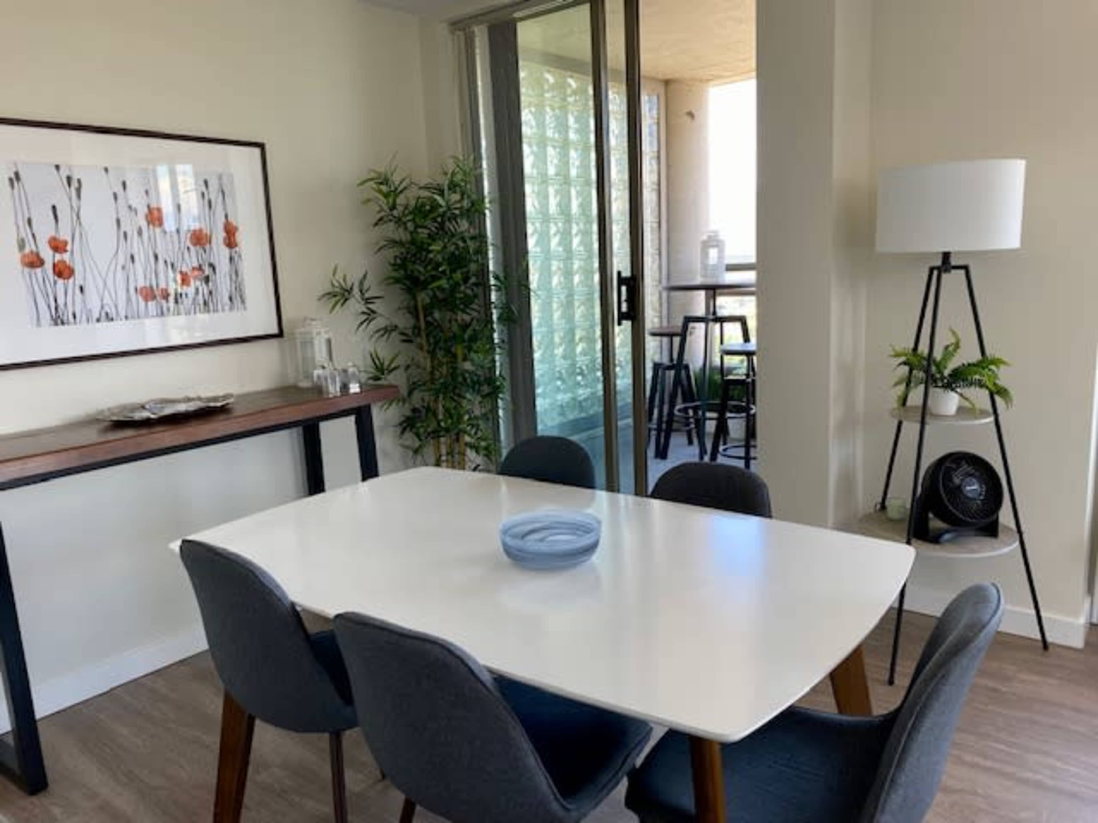 A dining area features a white table surrounded by gray chairs, with a large window and a balcony in the background.