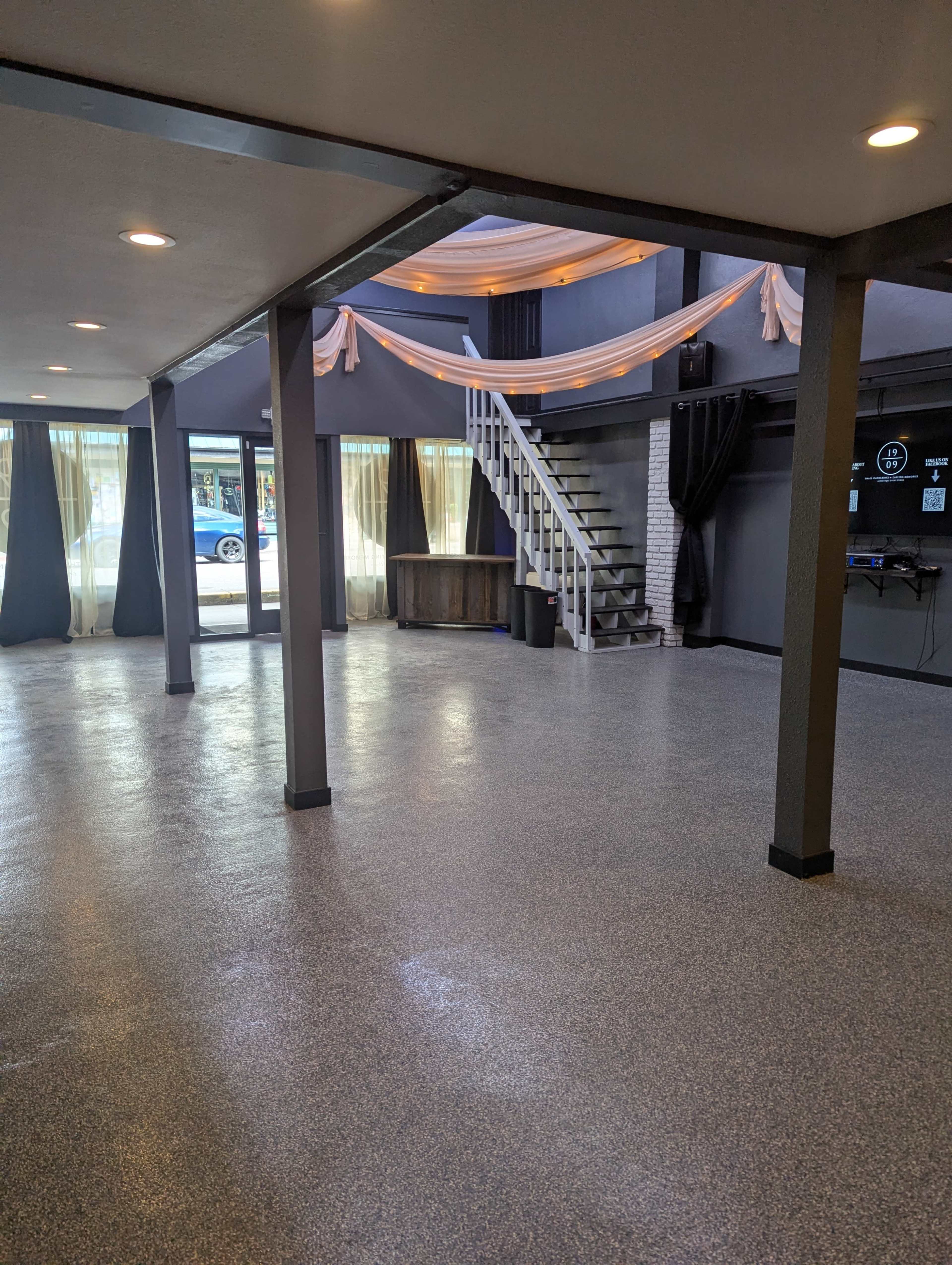 The image shows a spacious, empty interior room with polished flooring, large windows, and a staircase leading to an upper level.