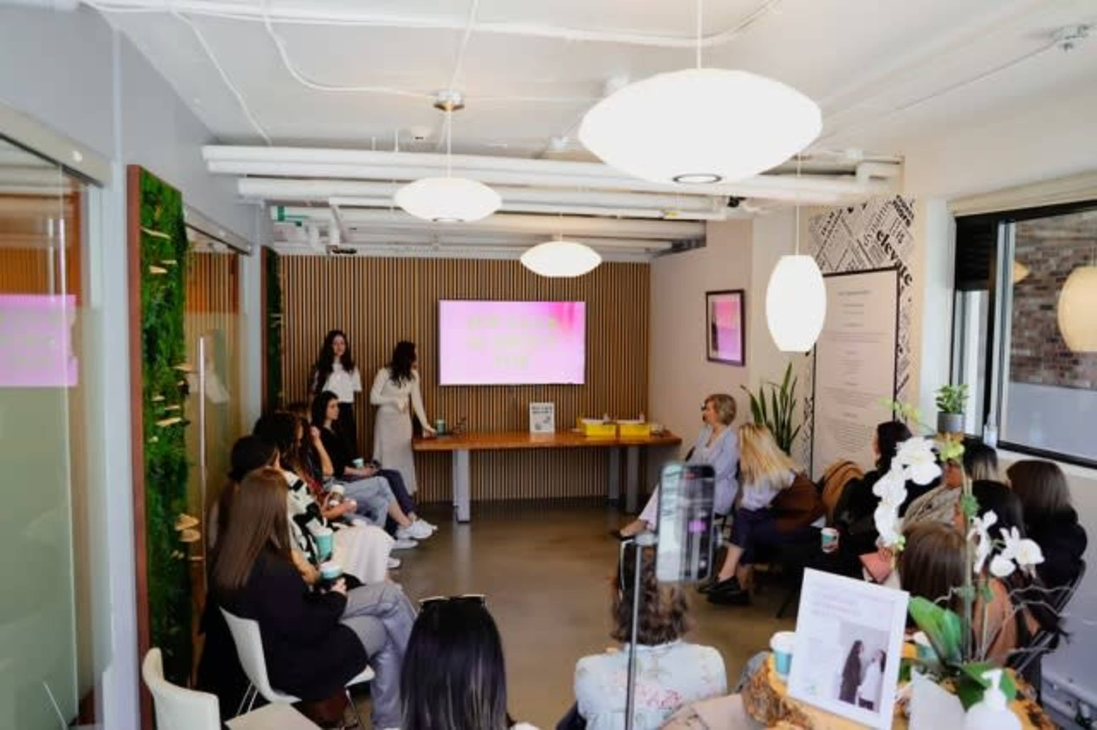 A group of people sits in a modern conference room, attentively watching a presentation on a screen while two presenters stand at the front.
