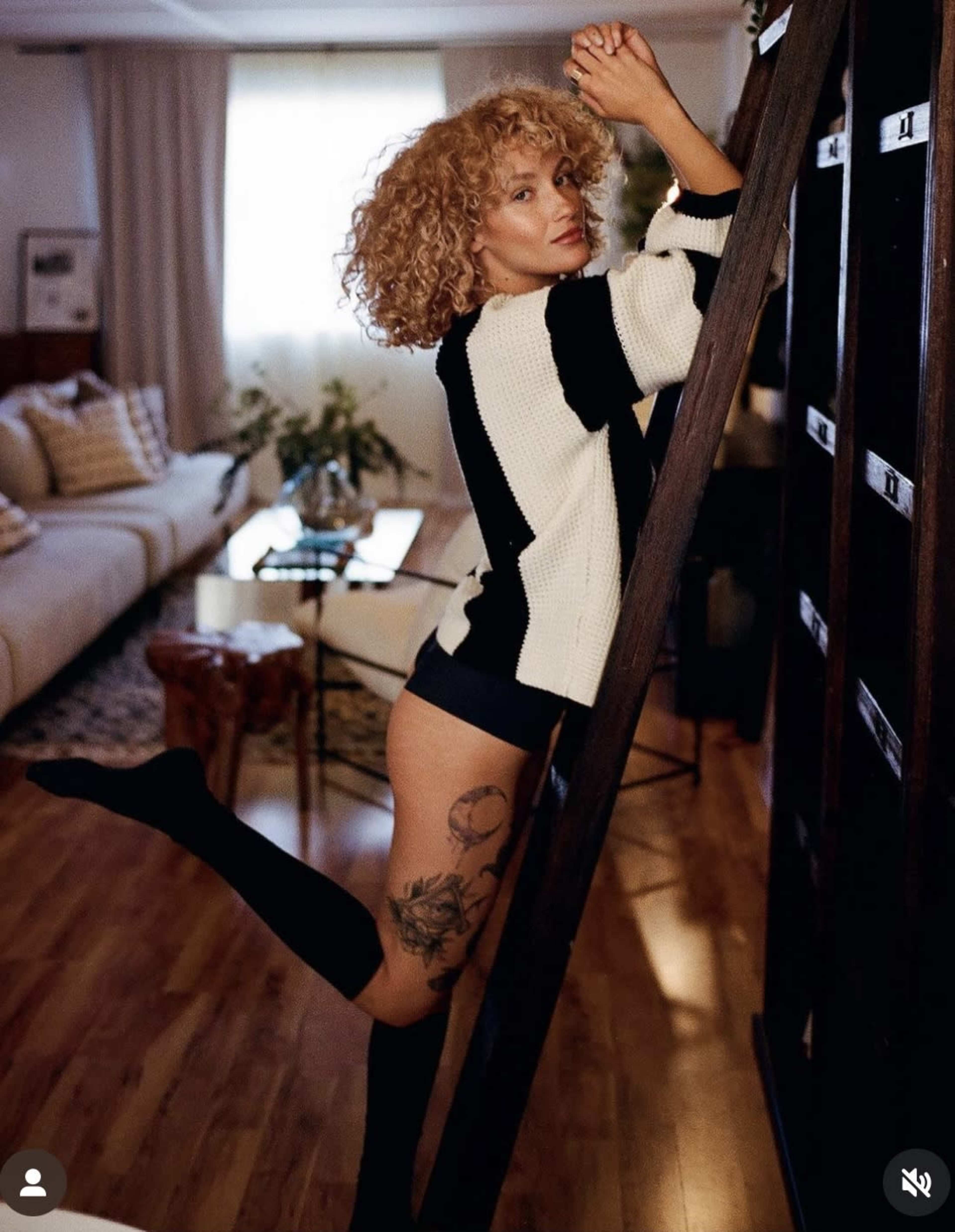 A person with curly hair poses on a wooden ladder in a well-lit room, showcasing a tattoo on their leg.