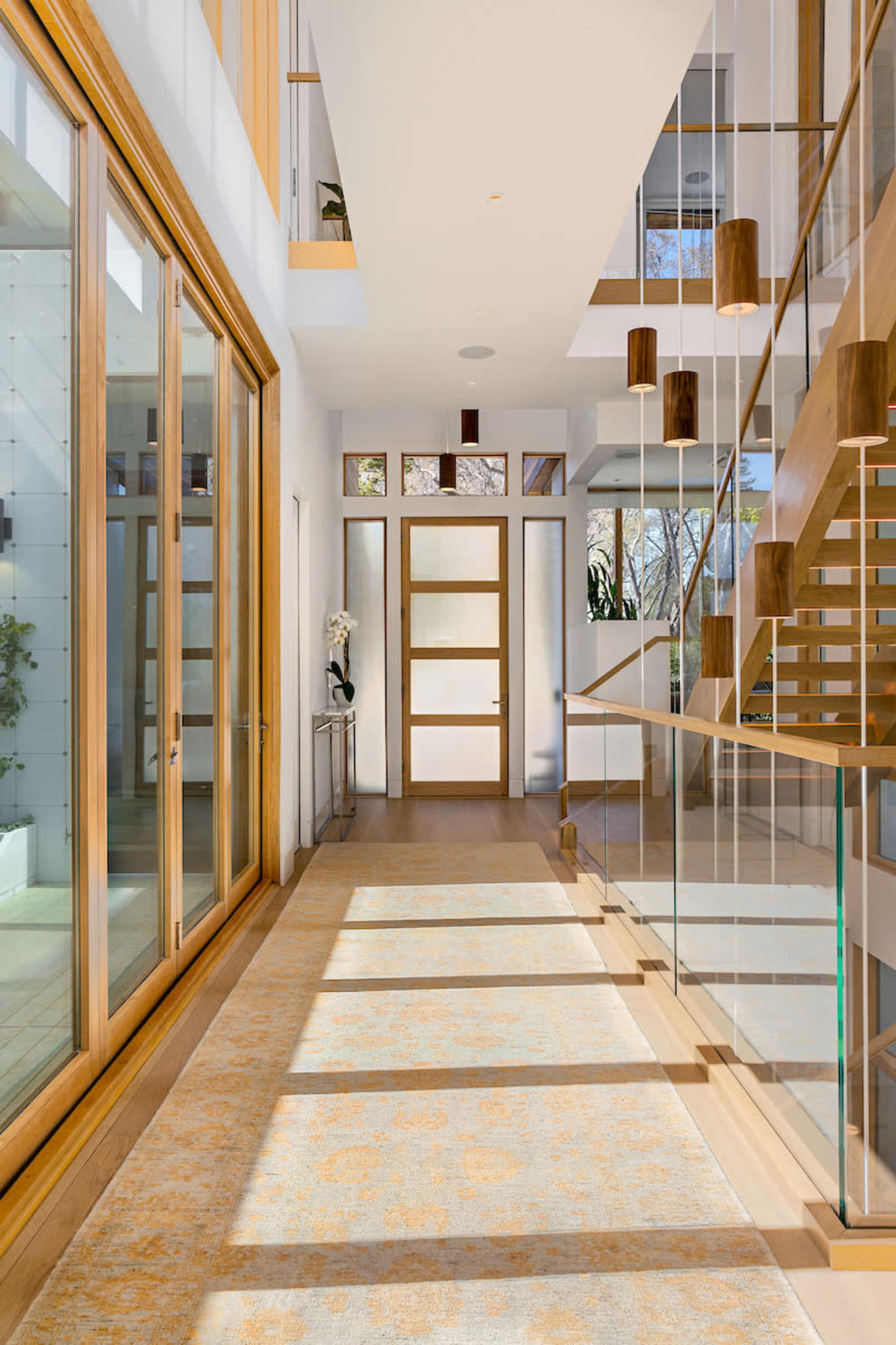 A modern hallway with large windows, a wooden staircase, and a patterned area rug.