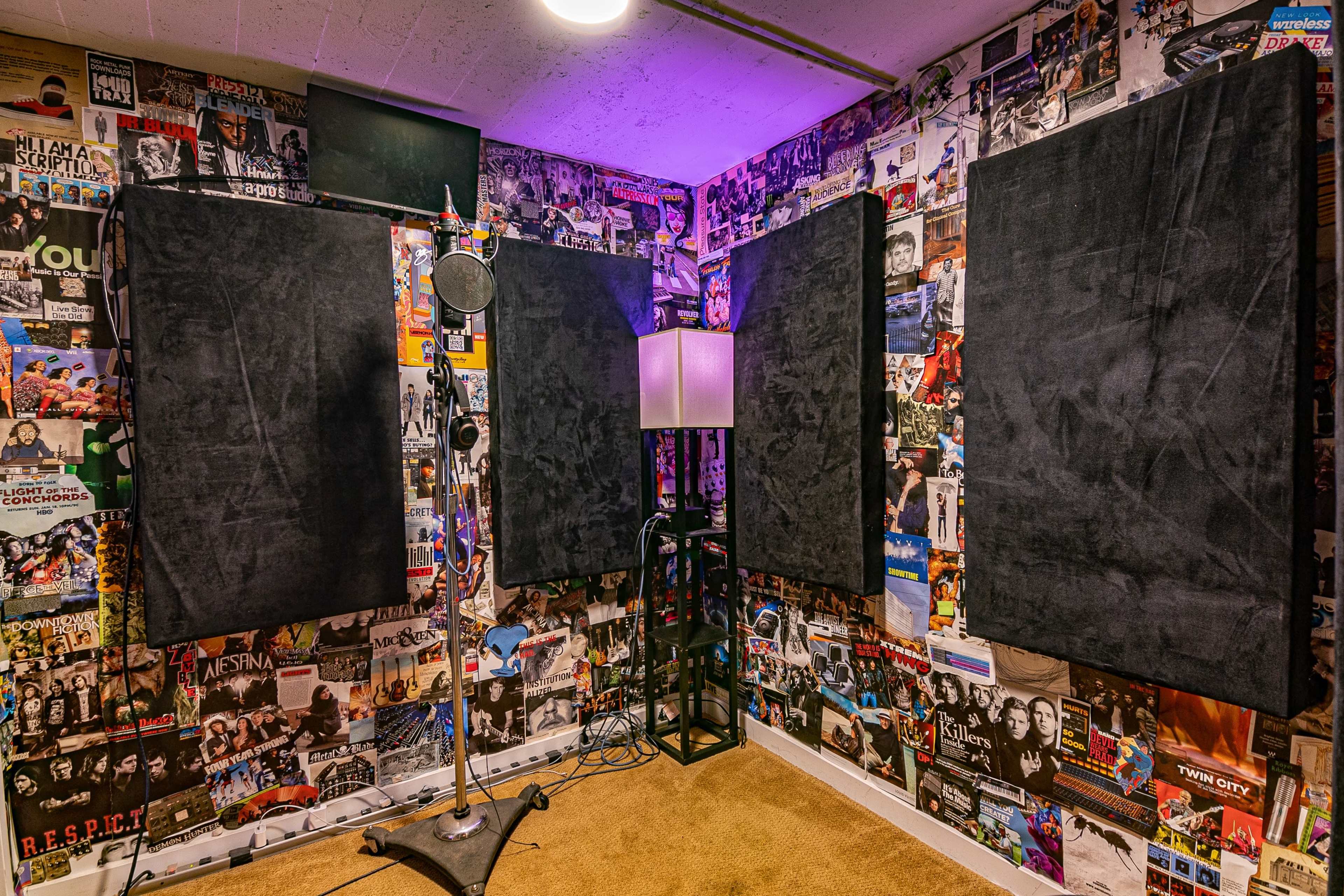 A small soundproof recording studio is adorned with a collage of posters on the walls, featuring a microphone and a lamp in the corner.