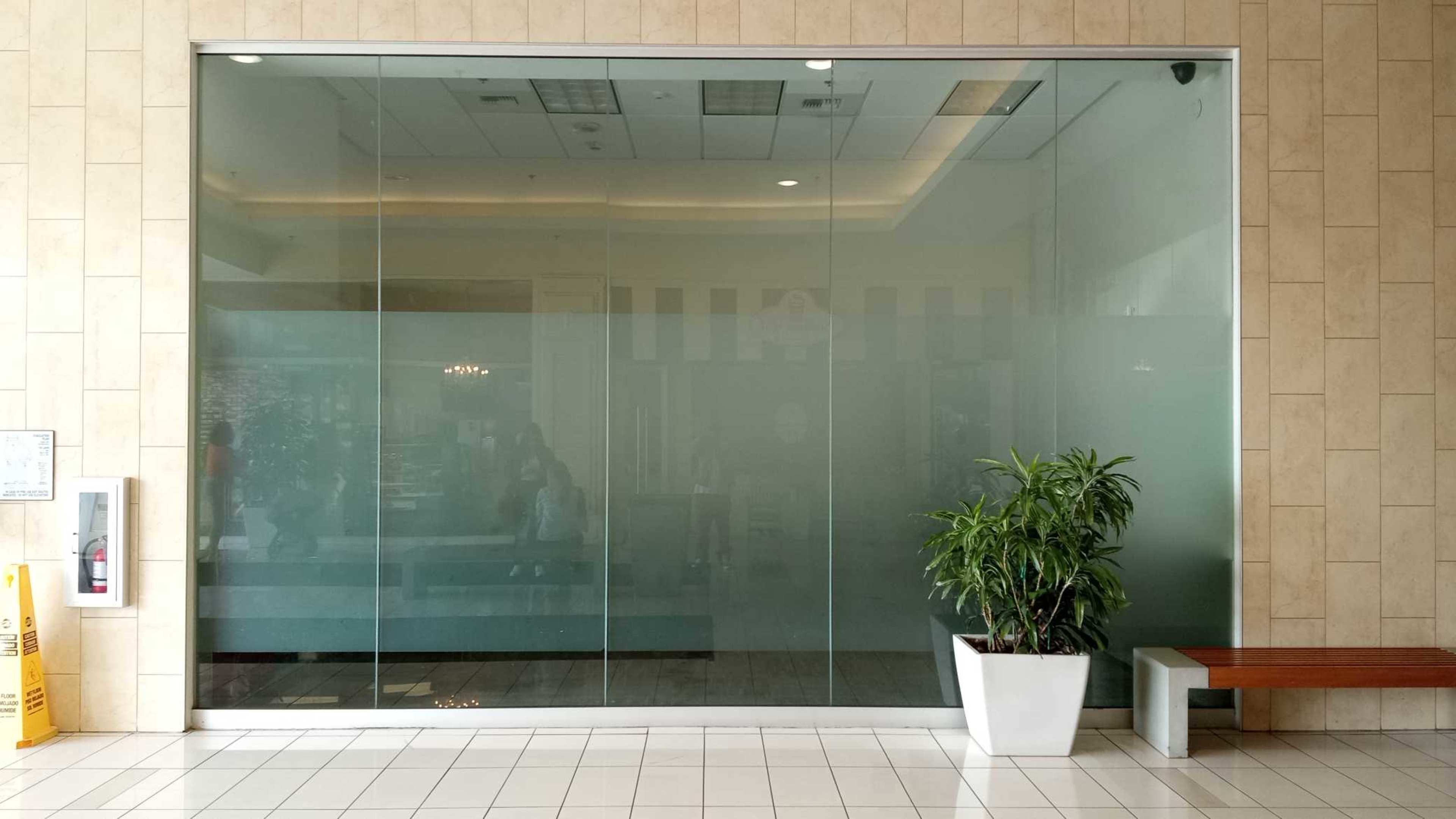 A large glass storefront with frosted sections and a potted plant beside a wooden bench in a mall.