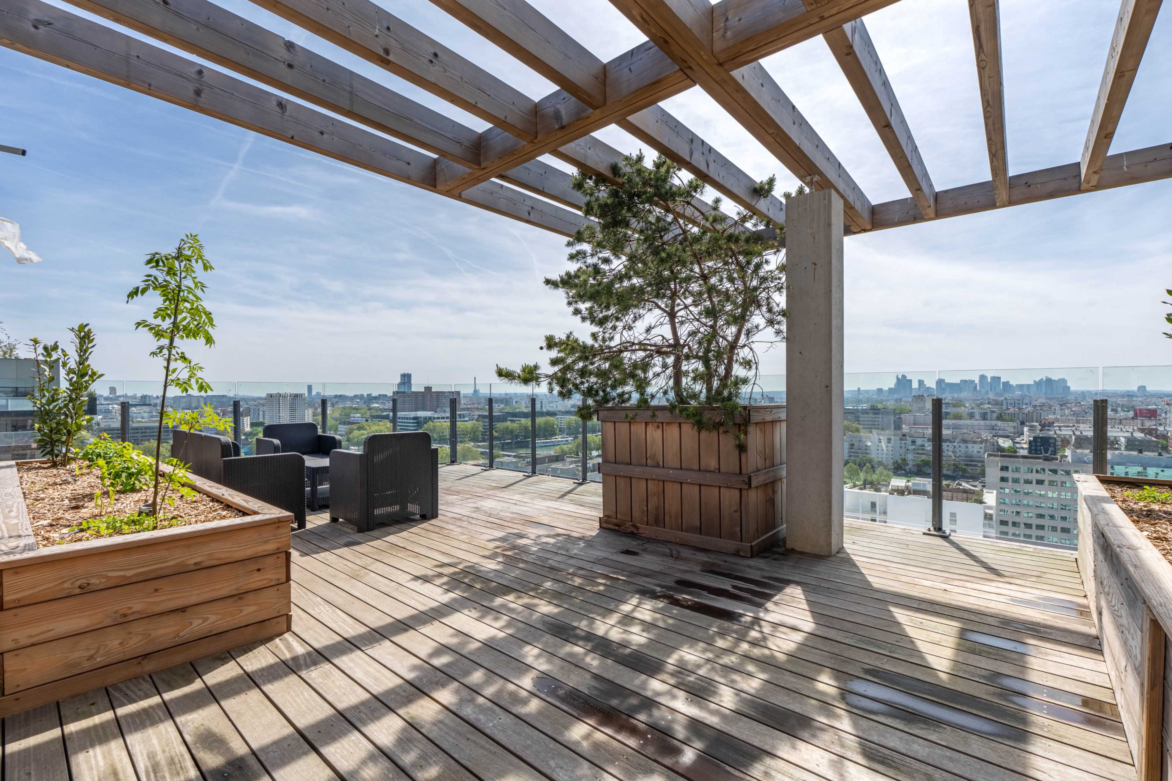 The image shows a rooftop terrace with wooden decking, planters, and seating, overlooking a city skyline under a clear sky.