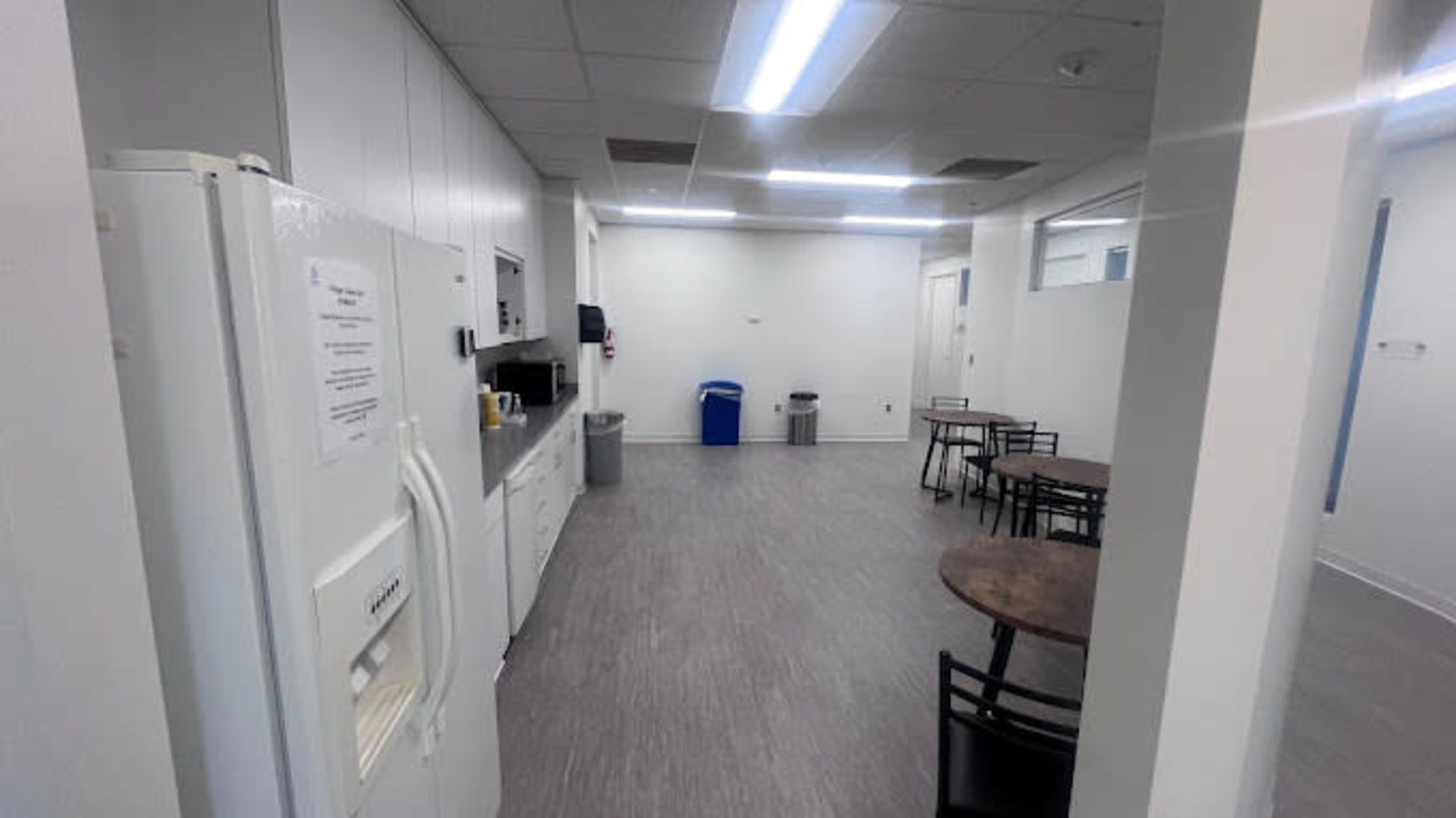 The image shows a spacious break room with a refrigerator, coffee machine, and multiple tables in a well-lit setting.