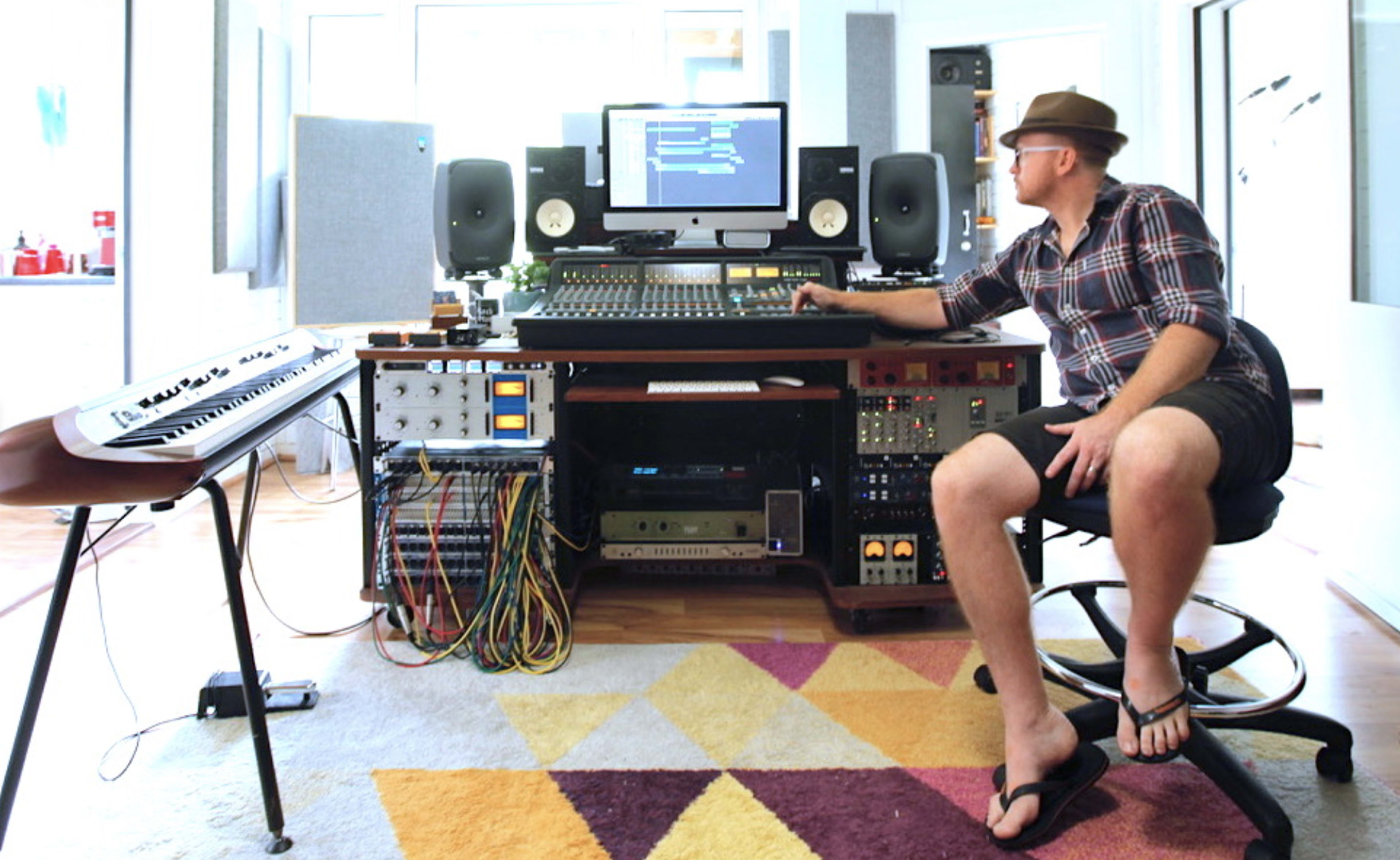 A person sits at a music production desk with various audio equipment and a computer screen in a well-lit studio.