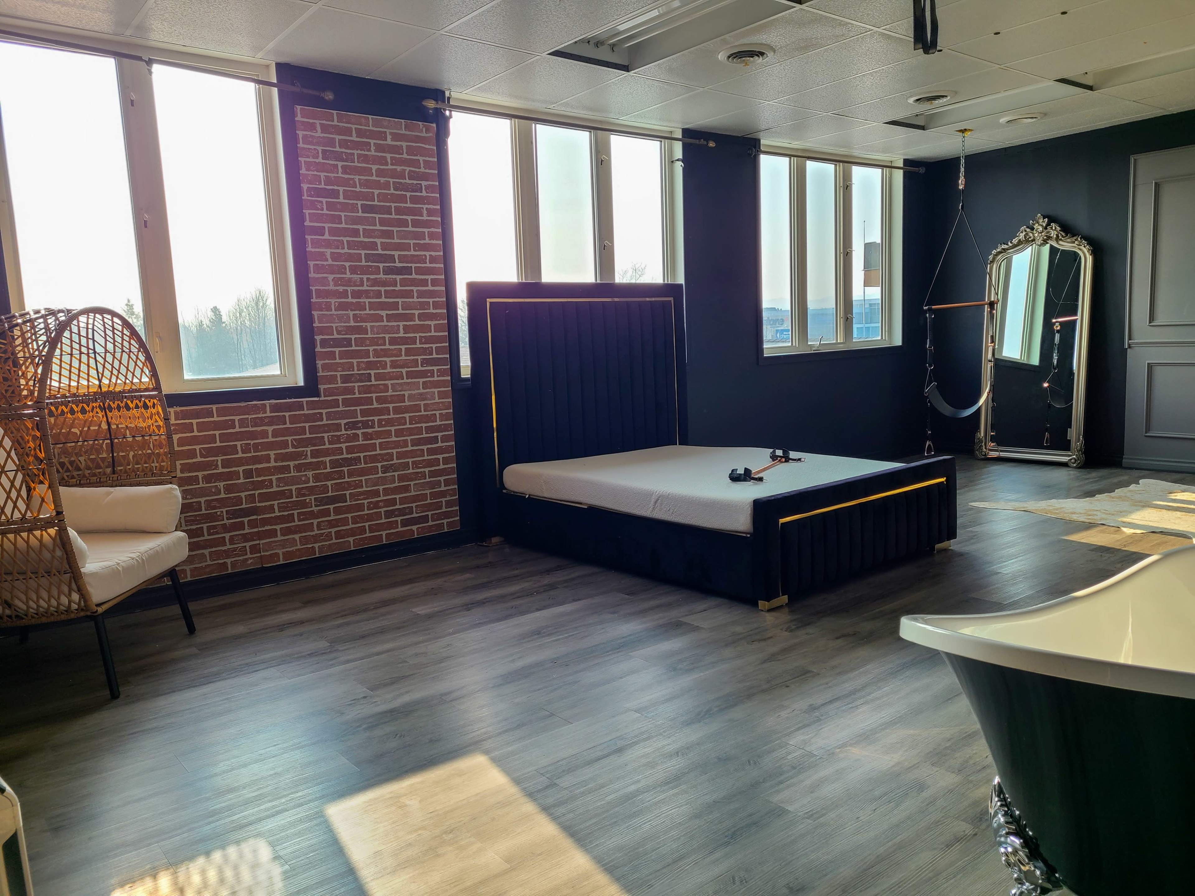 The room features a modern design with a bed, a vintage mirror, a freestanding bathtub, and a rattan chair against a brick wall.