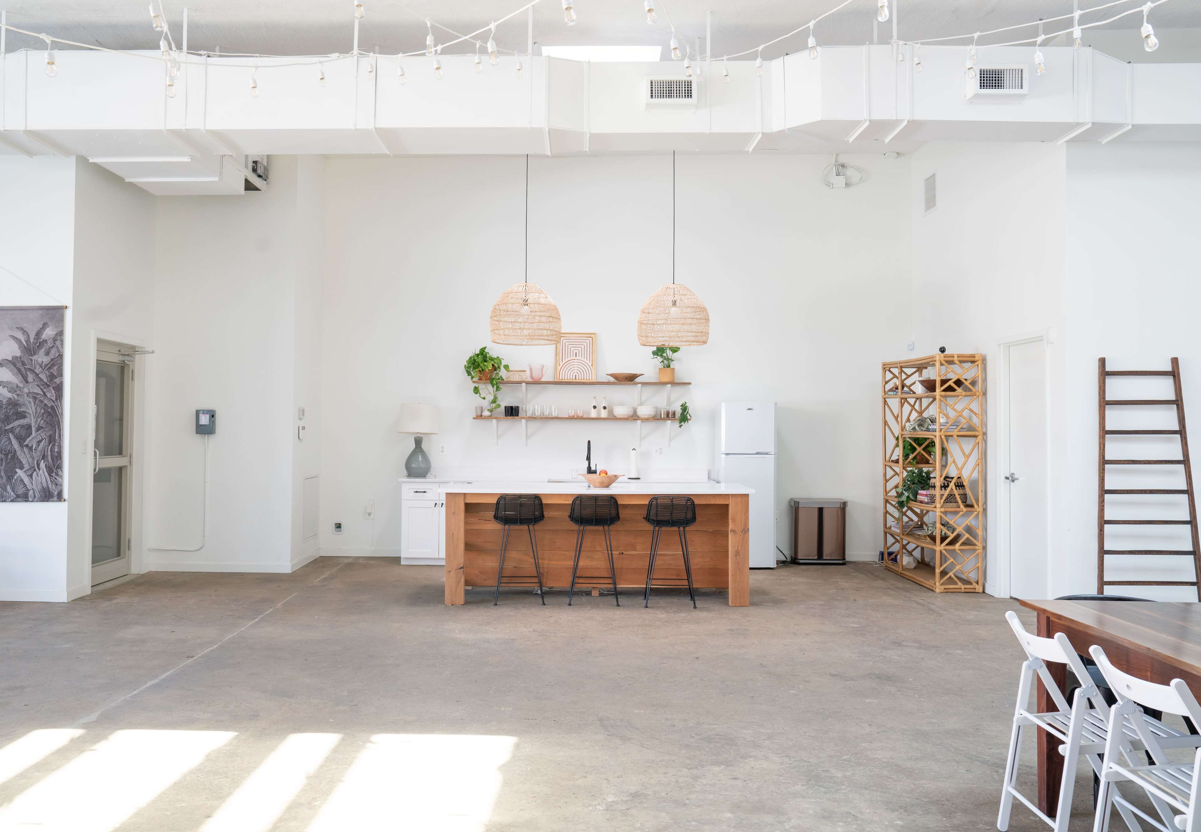 A spacious, modern kitchen with a wooden island, three black bar stools, and pendant lighting above, complemented by open shelving and a refrigerator in a light-filled environment.