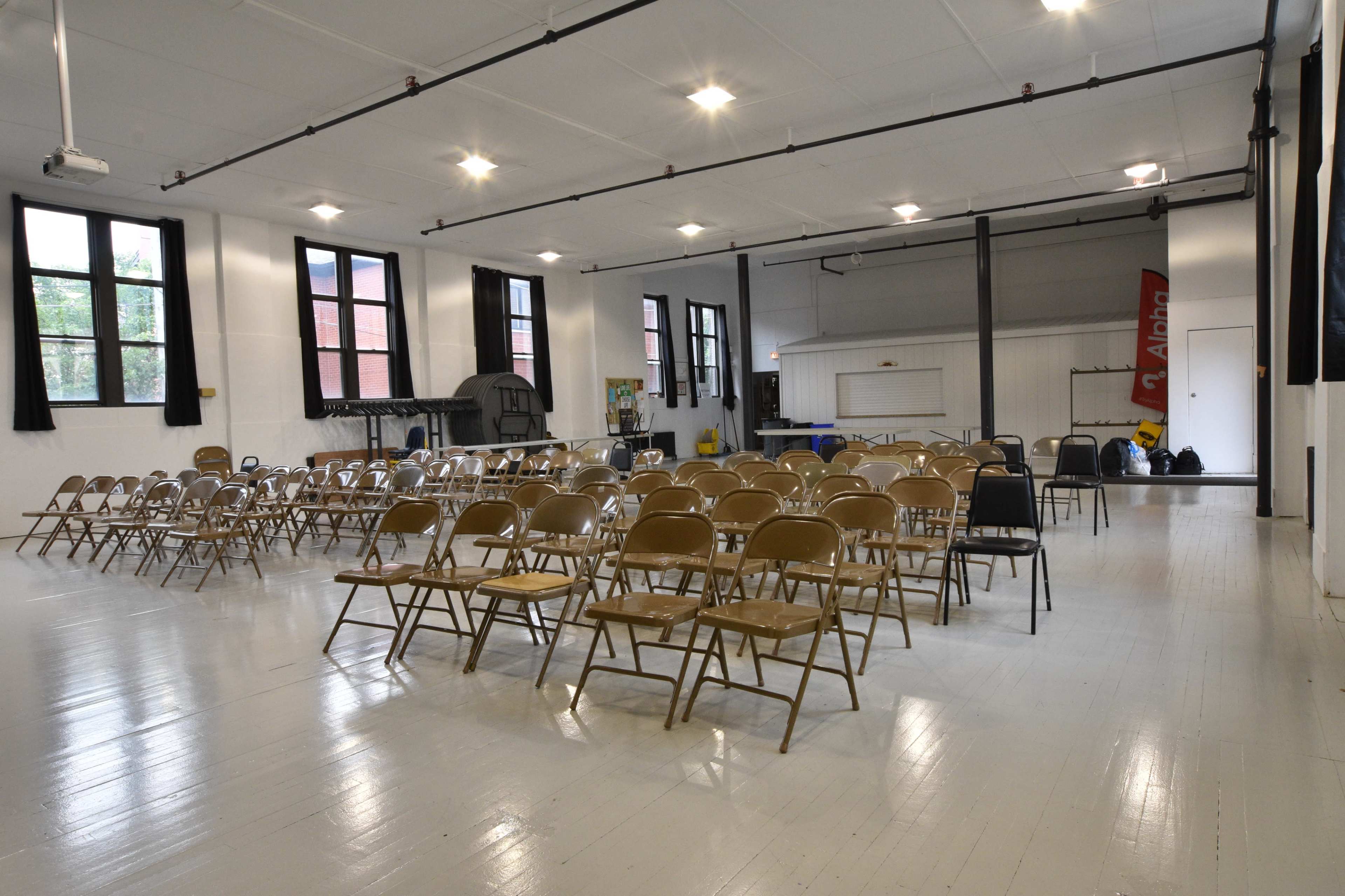 A spacious room with rows of folded metal chairs arranged facing a blank wall, illuminated by overhead lights.