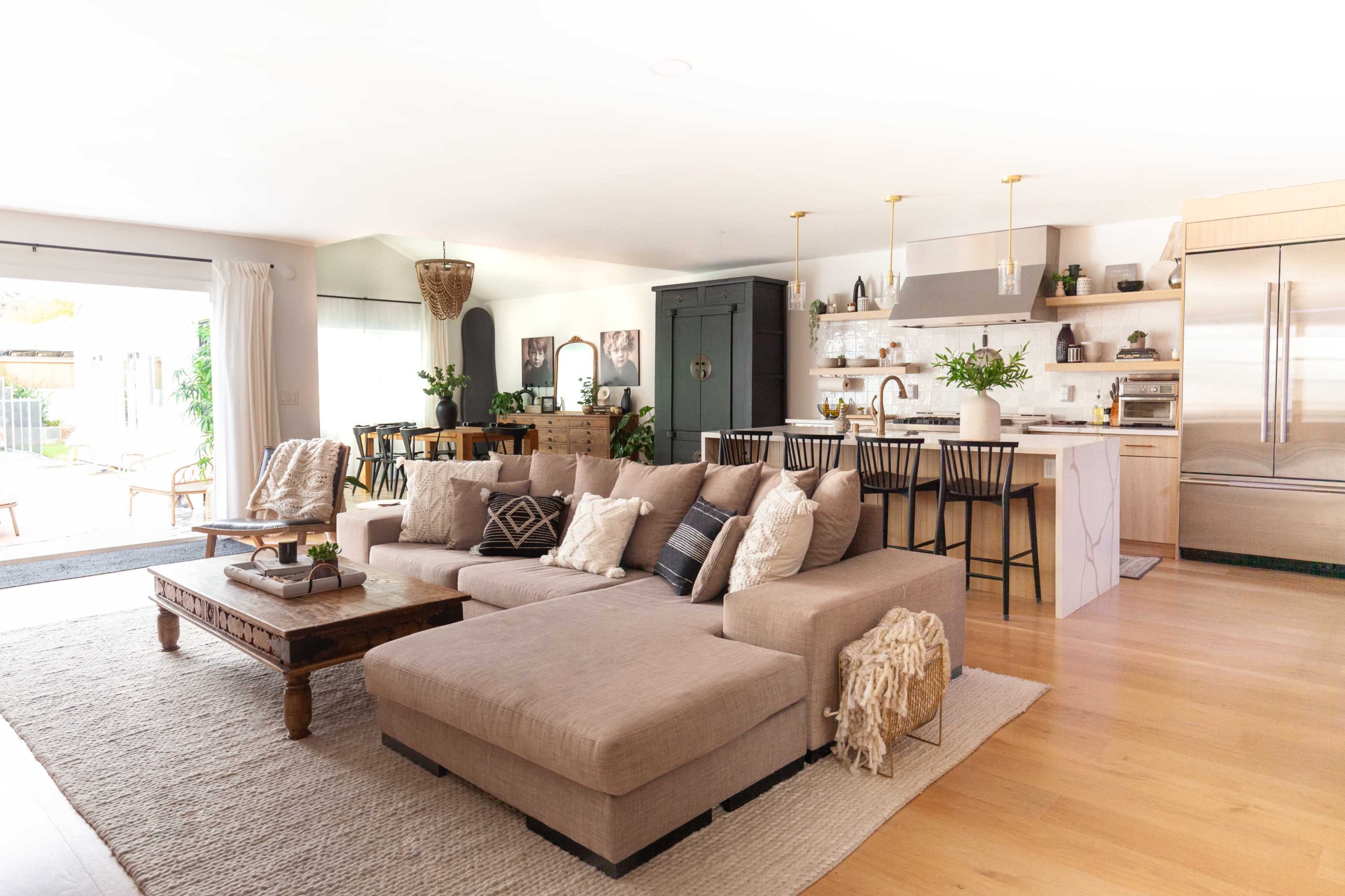A modern living room with a large beige sectional sofa, a wooden coffee table, and an open kitchen featuring bar stools and a sleek refrigerator.
