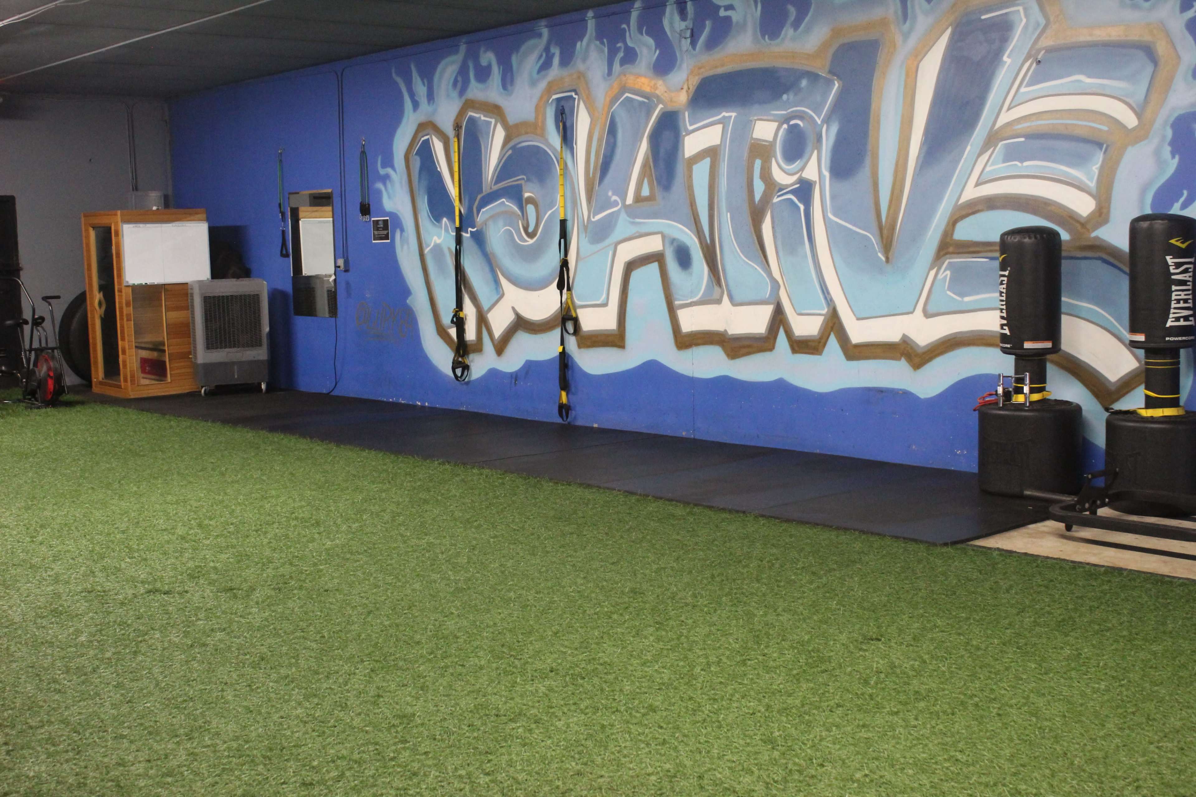 A gym space with artificial turf, a blue wall featuring graffiti that says "Volative," and fitness equipment including punching bags and suspension training straps.