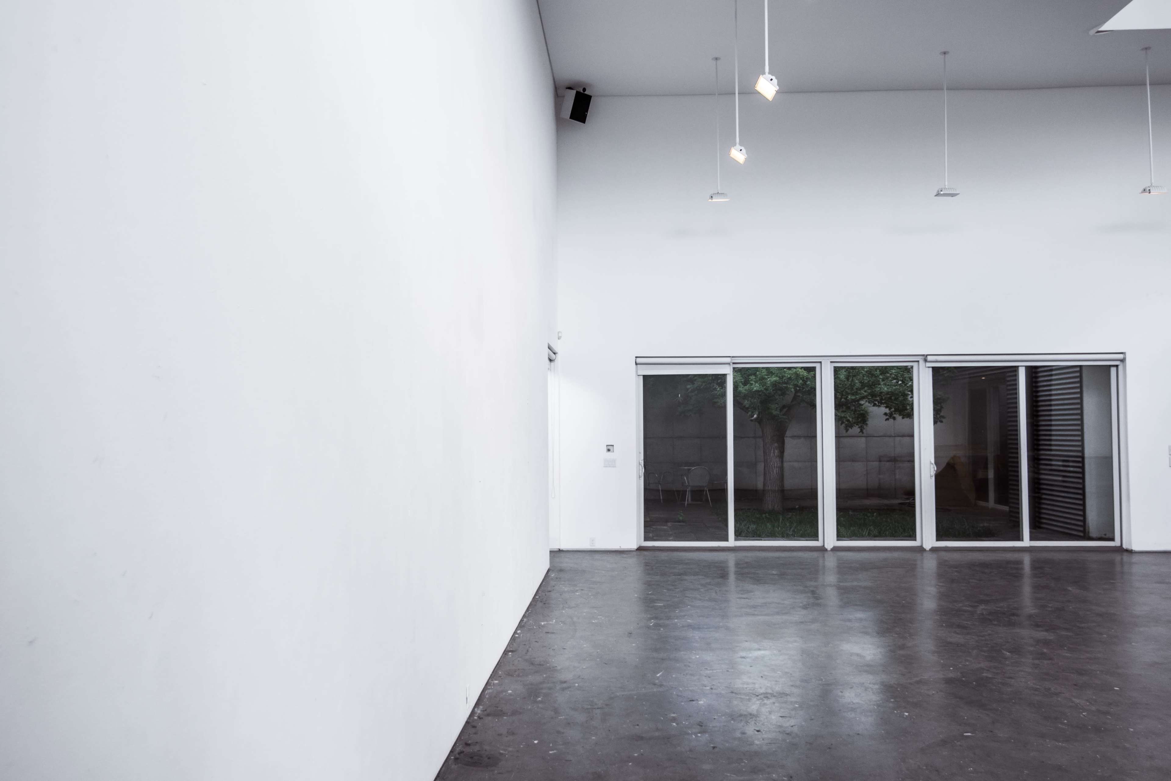 The image shows a minimalist interior space with a large white wall and sliding glass doors leading to an outdoor area with visible greenery.