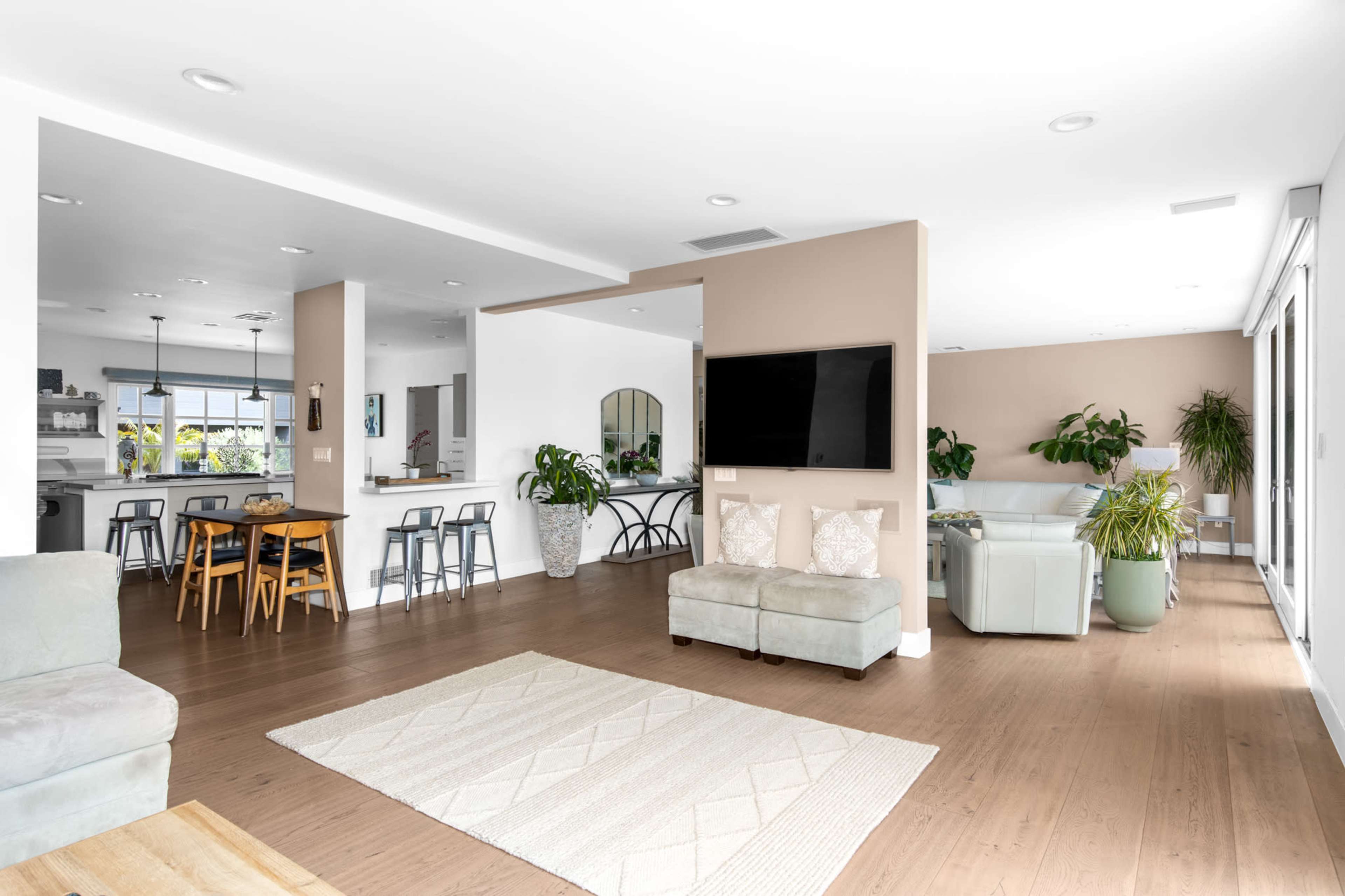 The image shows a modern open-concept living space that includes a seating area, dining table, kitchen bar, and large windows with plants integrated into the decor.