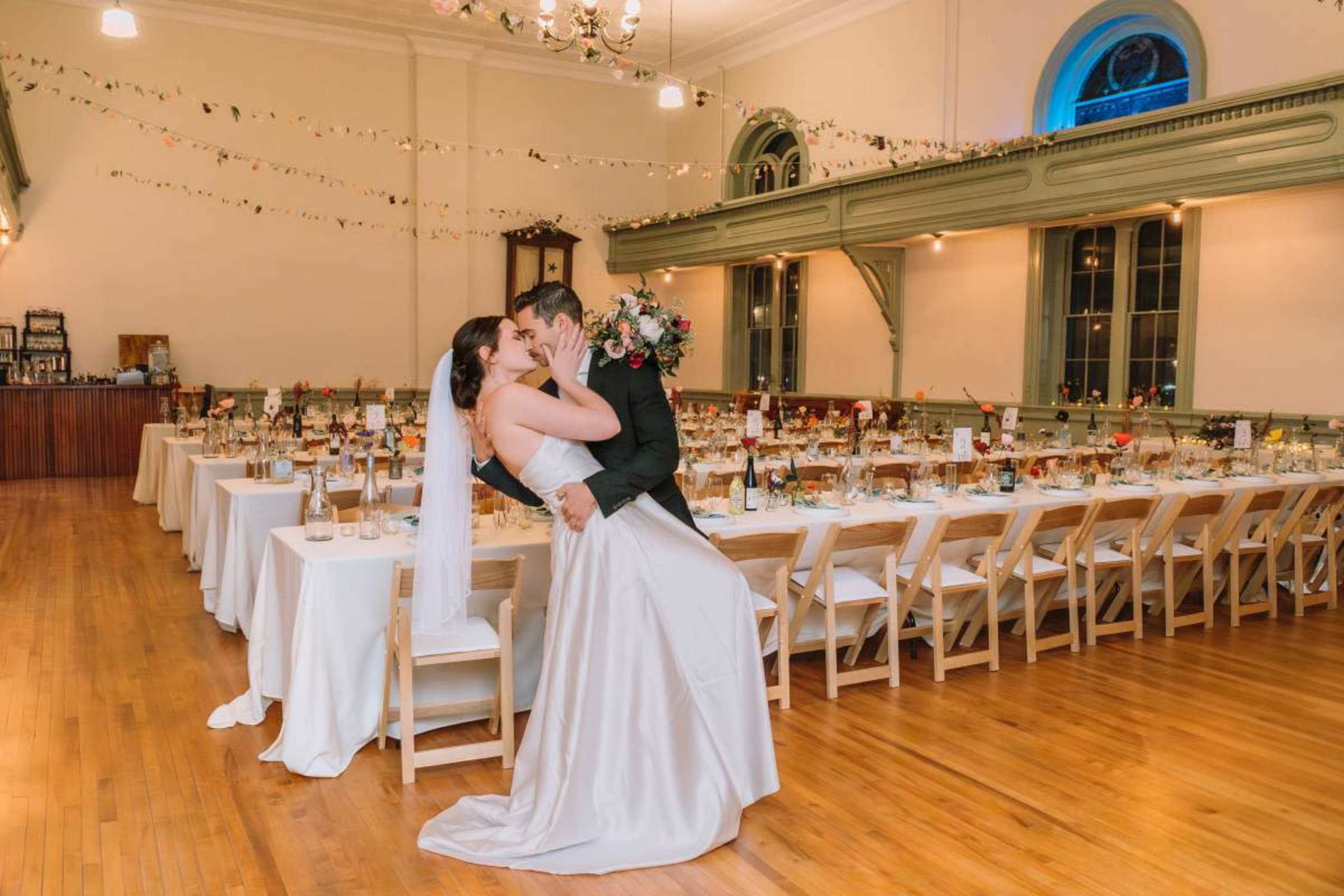 A bride and groom kiss in a decorated reception hall, surrounded by tables set for a wedding dinner.
