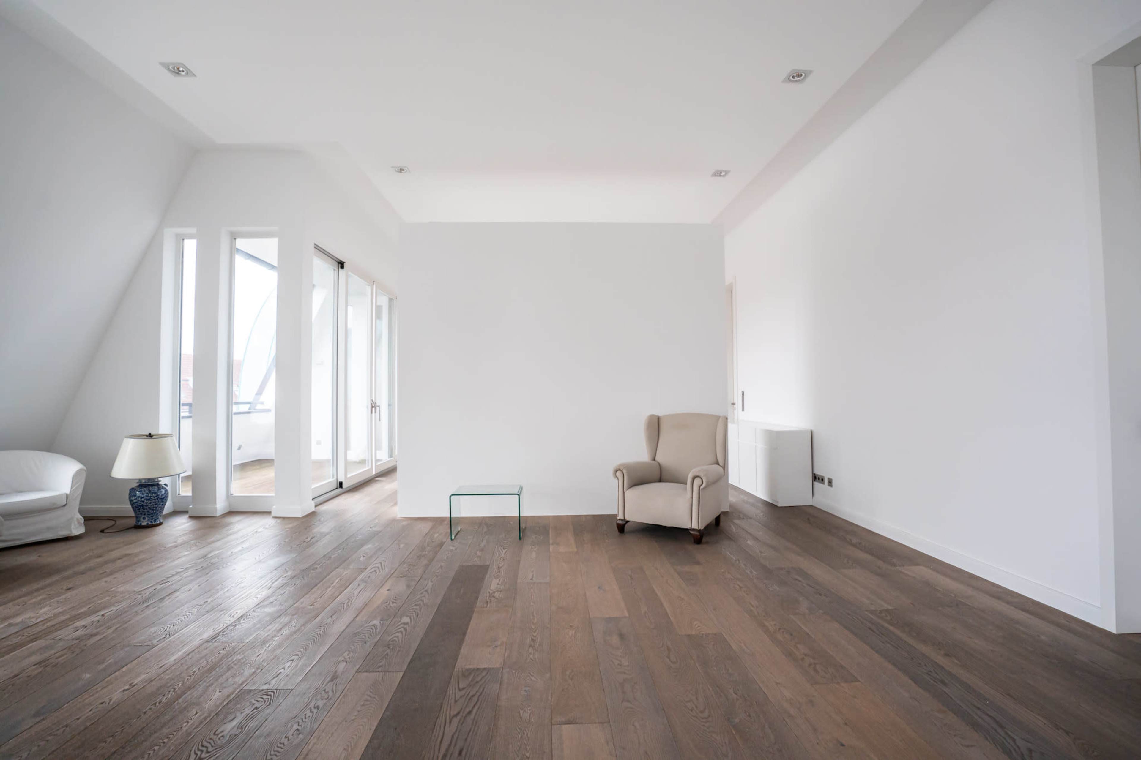The image shows a spacious, minimally furnished room with hardwood flooring, a single armchair, a glass table, and large windows allowing natural light.
